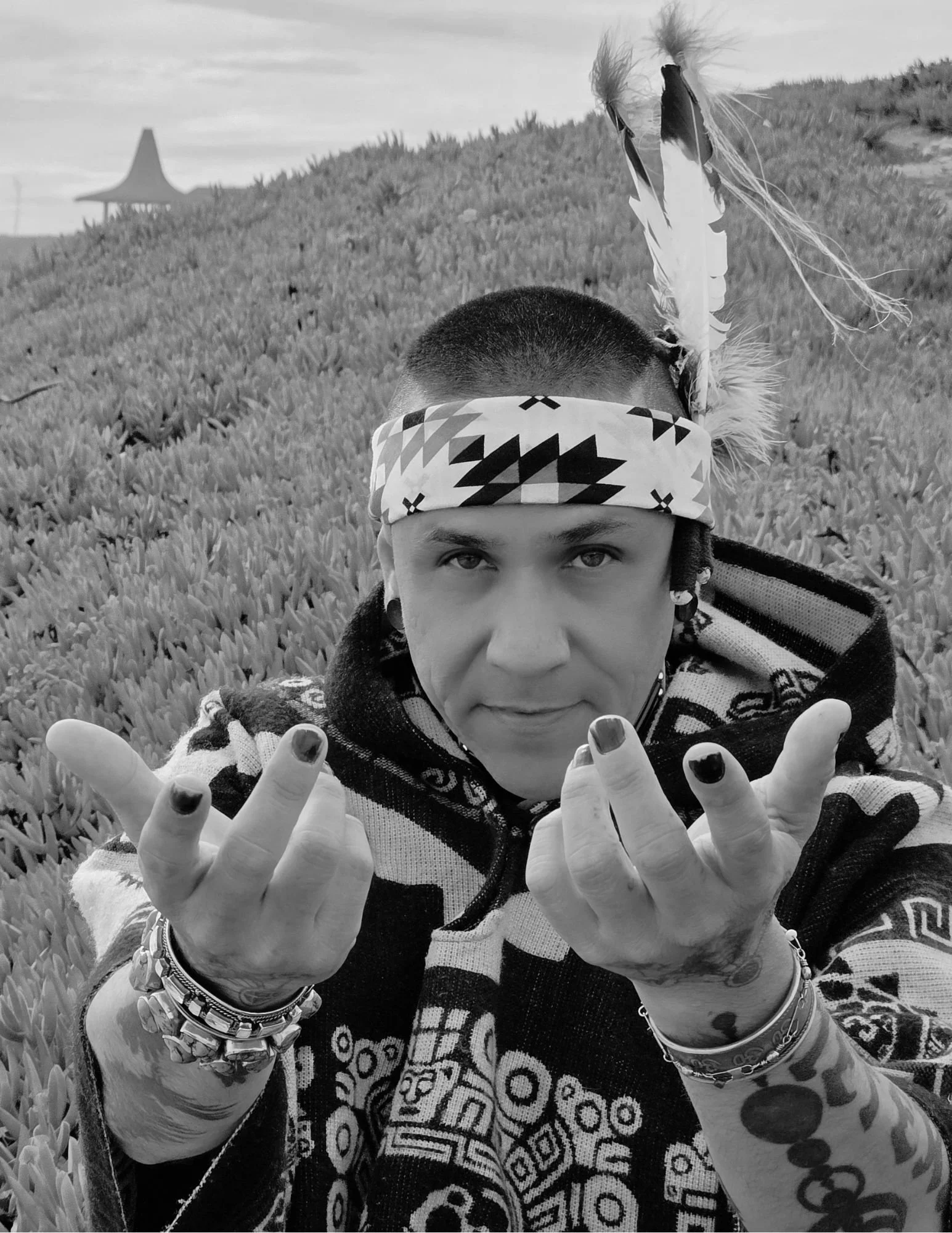 A person with short hair wearing a patterned headband and a jacket with tribal and geometric designs, standing in a field of plants, with a mountain and a tent-like structure in the background. The person is holding their hands up with fingernails painted dark.