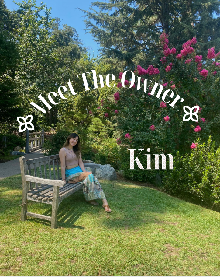 A woman sitting on a wooden bench in a lush garden with green trees and pink flowers, smiling at the camera, with overlay text 'Meet The Owner Kim'.