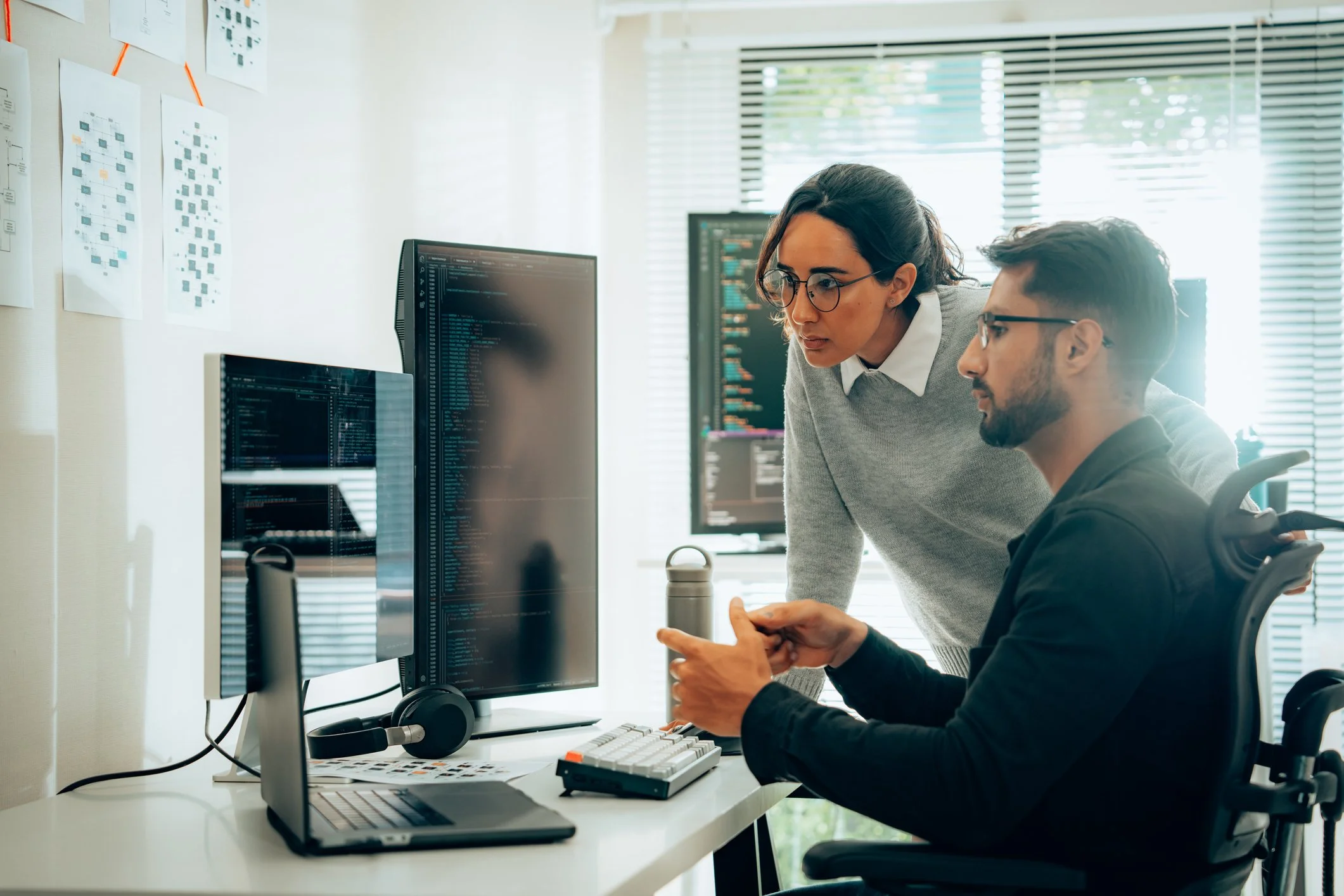 Dos personas trabajando en una oficina, una está sentada en una silla de oficina con una computadora y otra está de pie, mirando la pantalla, con más monitores en el fondo y documentos en la pared.