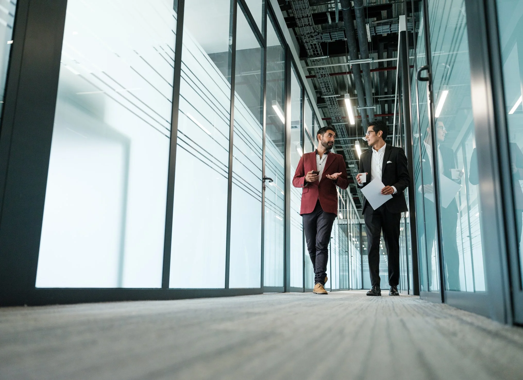 Dos hombres de negocios conversando y caminando por un pasillo de oficina moderna con paredes de cristal y ambiente bien iluminado.