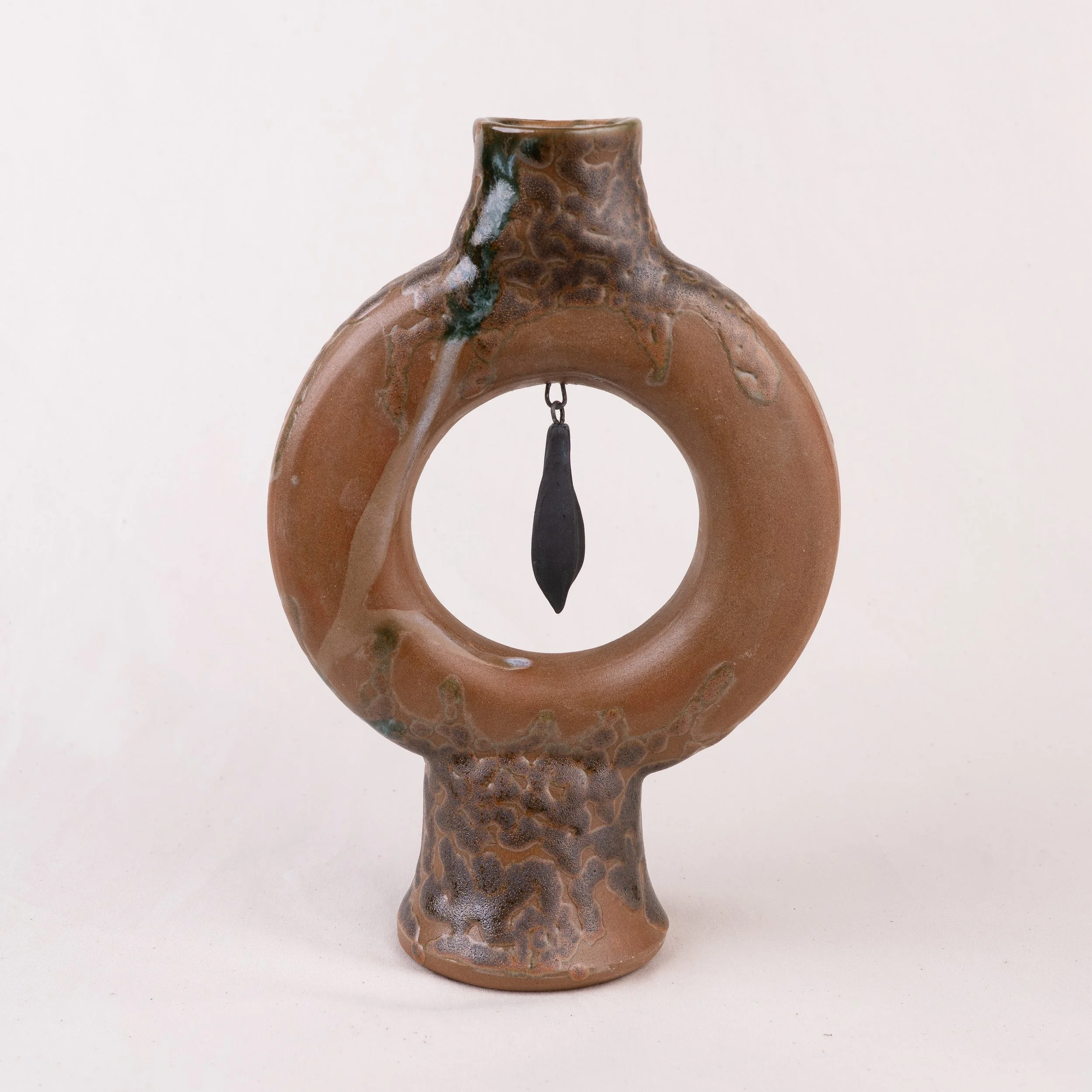 A ceramic sculptural vase resembling a ringed planet or a hollow ring with a small chain hanging inside, featuring a textured, mottled brown surface with darker and lighter patches.