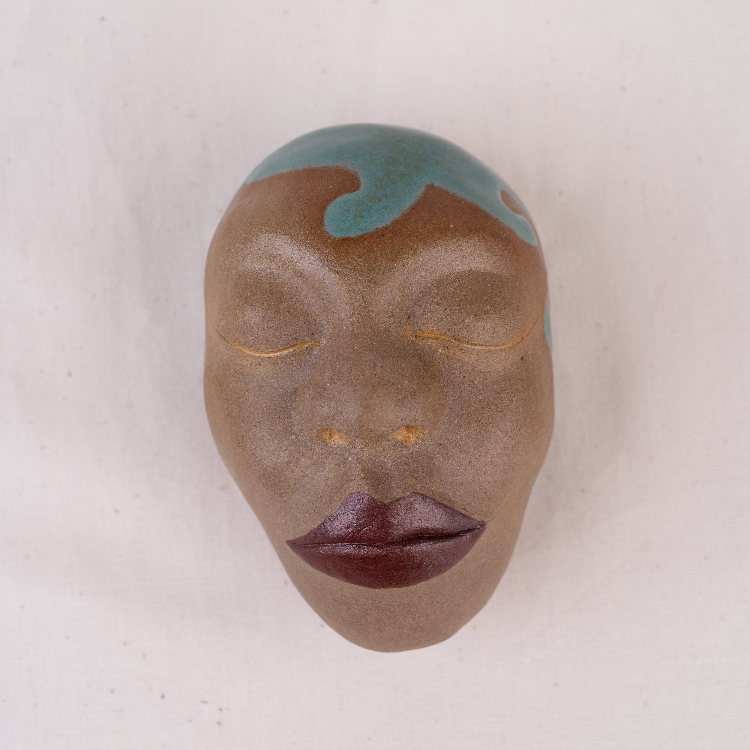 Close-up of a small face sculpture, featuring a combination of pink, blue, and gold colors, against a plain, light-colored background.