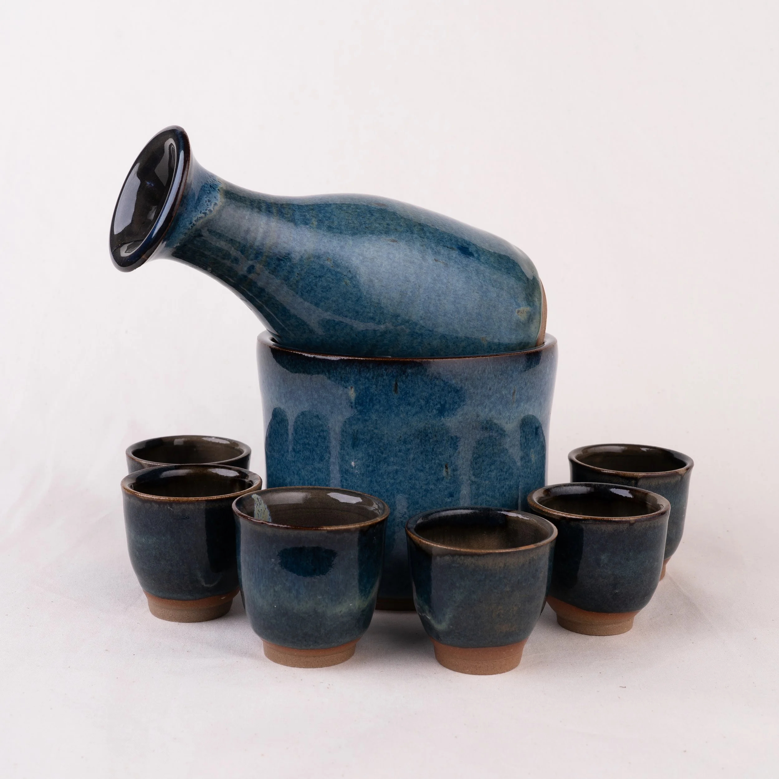 A ceramic sake set featuring a bottle, chilling bowl, and six cups with blue glaze finish, arranged on a white background.