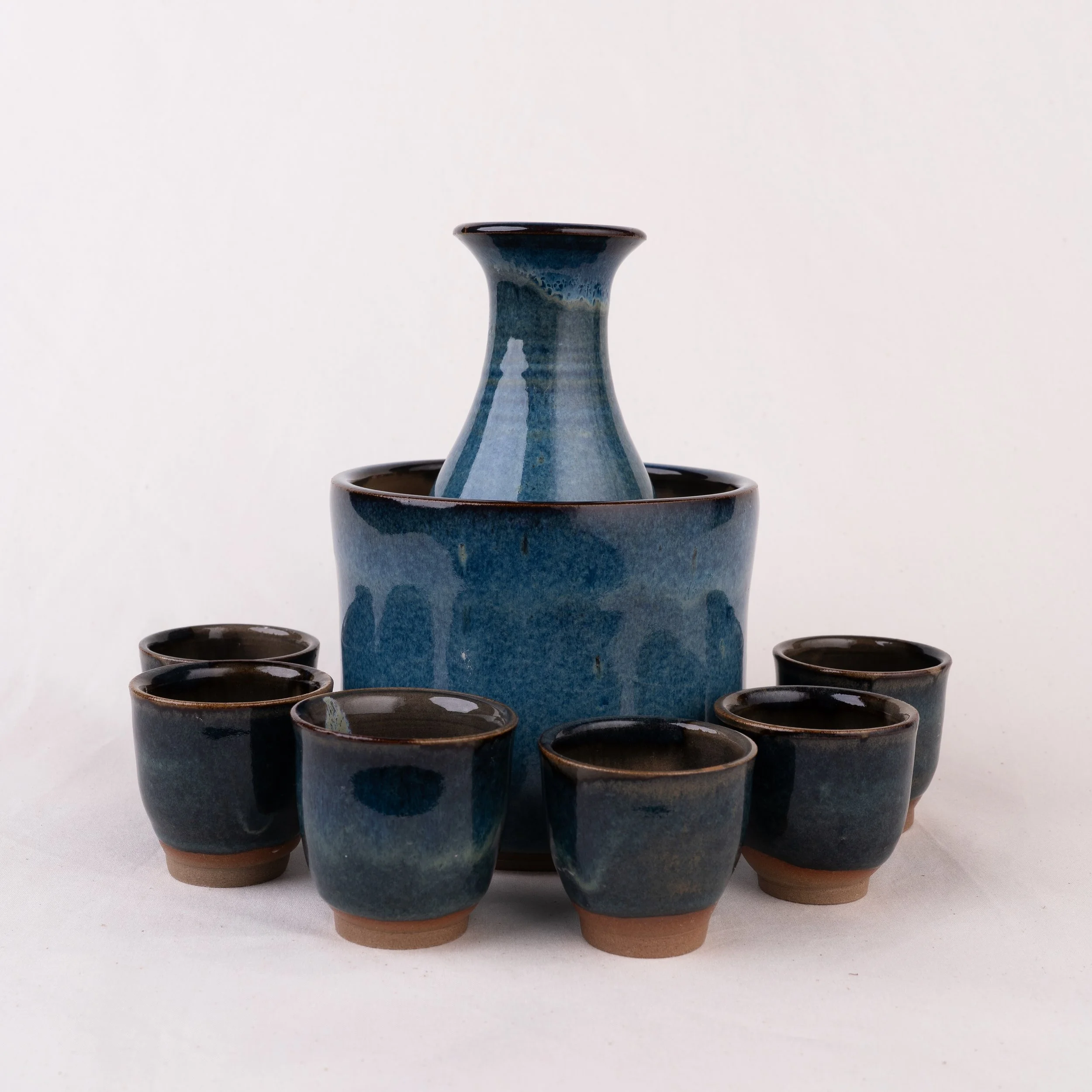 A ceramic sake set featuring a bottle, chilling bowl, and six cups with blue glaze finish, displayed on a white background.