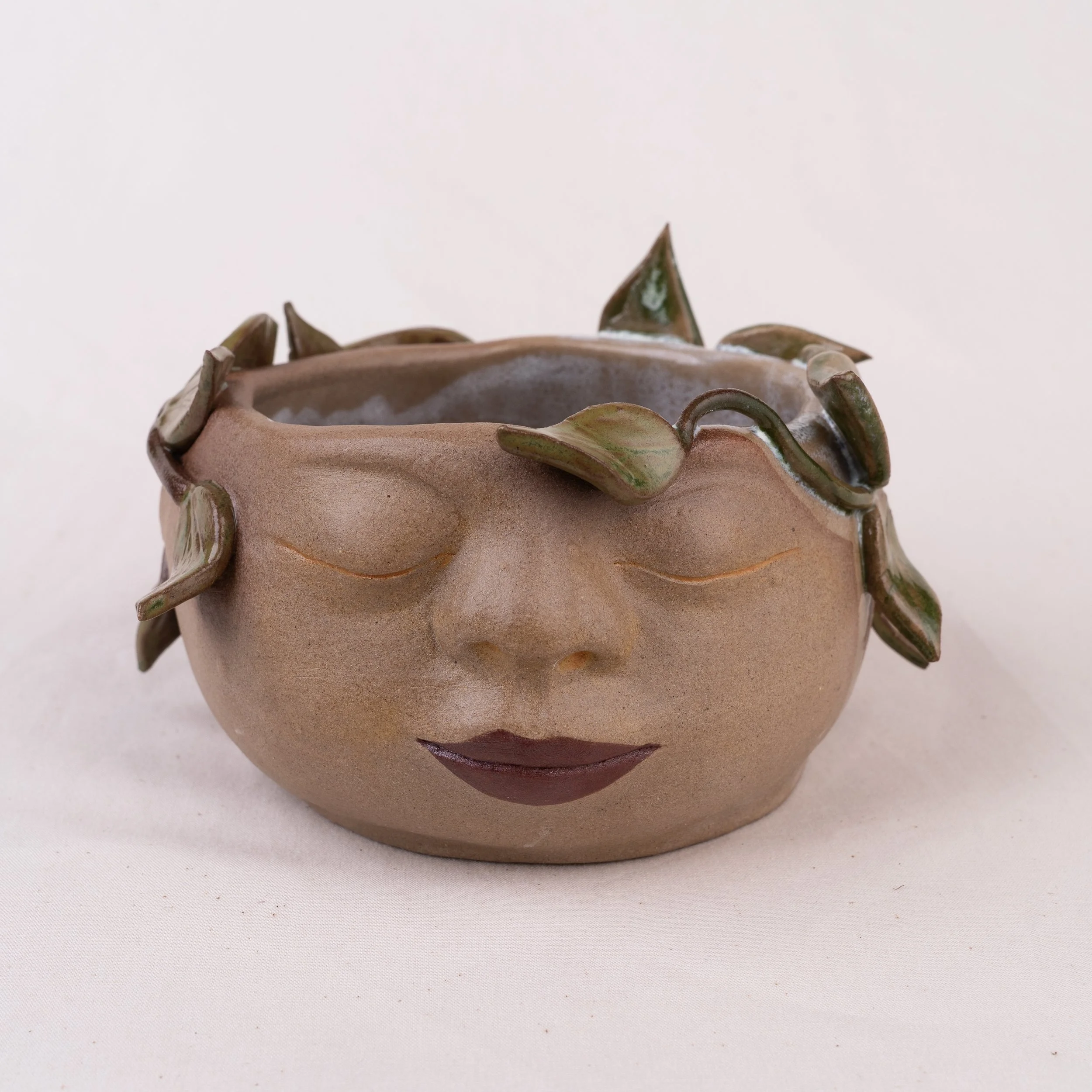 Close-up of a ceramic face shaped planter with a matte finish, featuring decorative leaves around its rim, set against a plain white background.