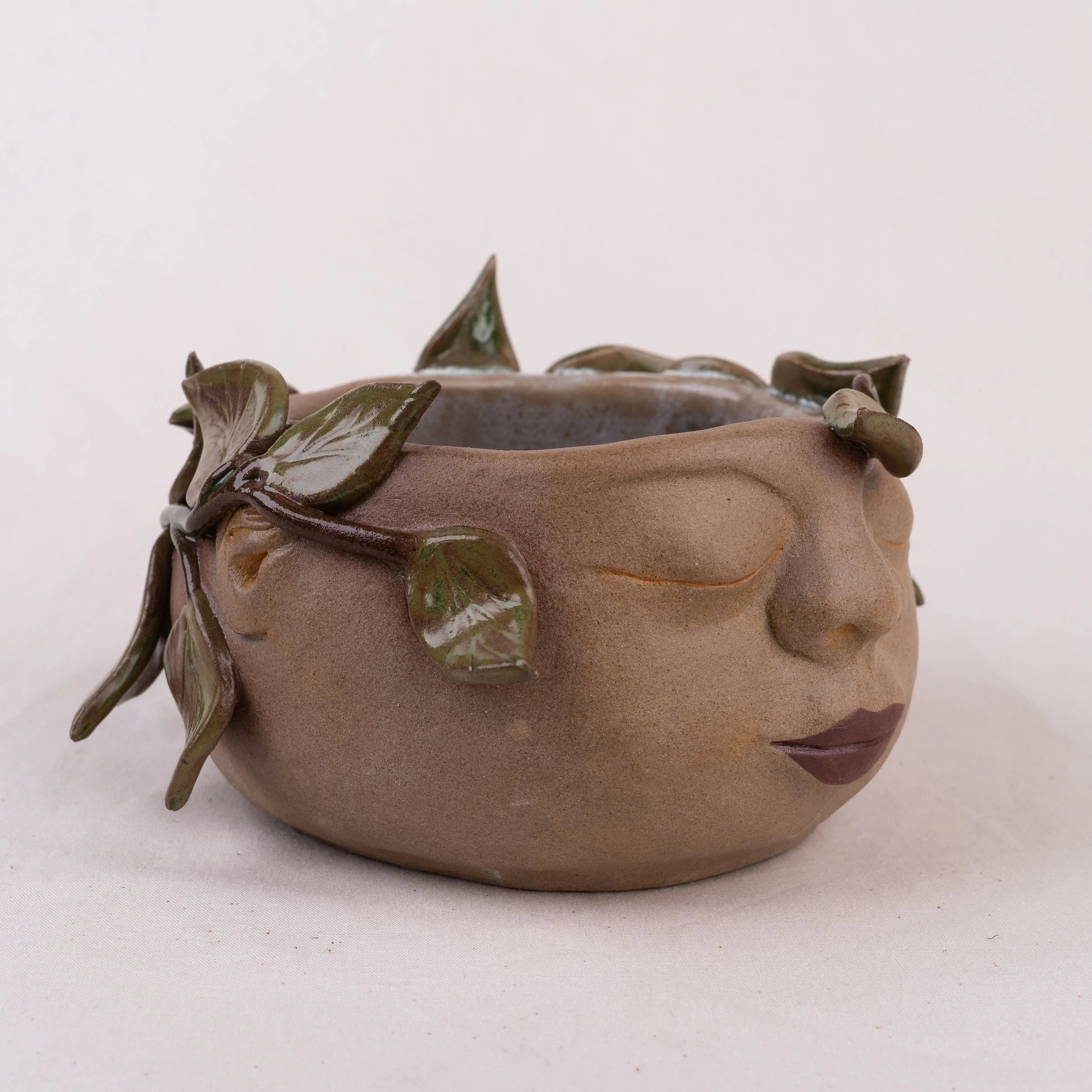 Close-up side view of a ceramic face shaped planter with green leafy details around the top edge, placed on a light-colored surface against a plain background.