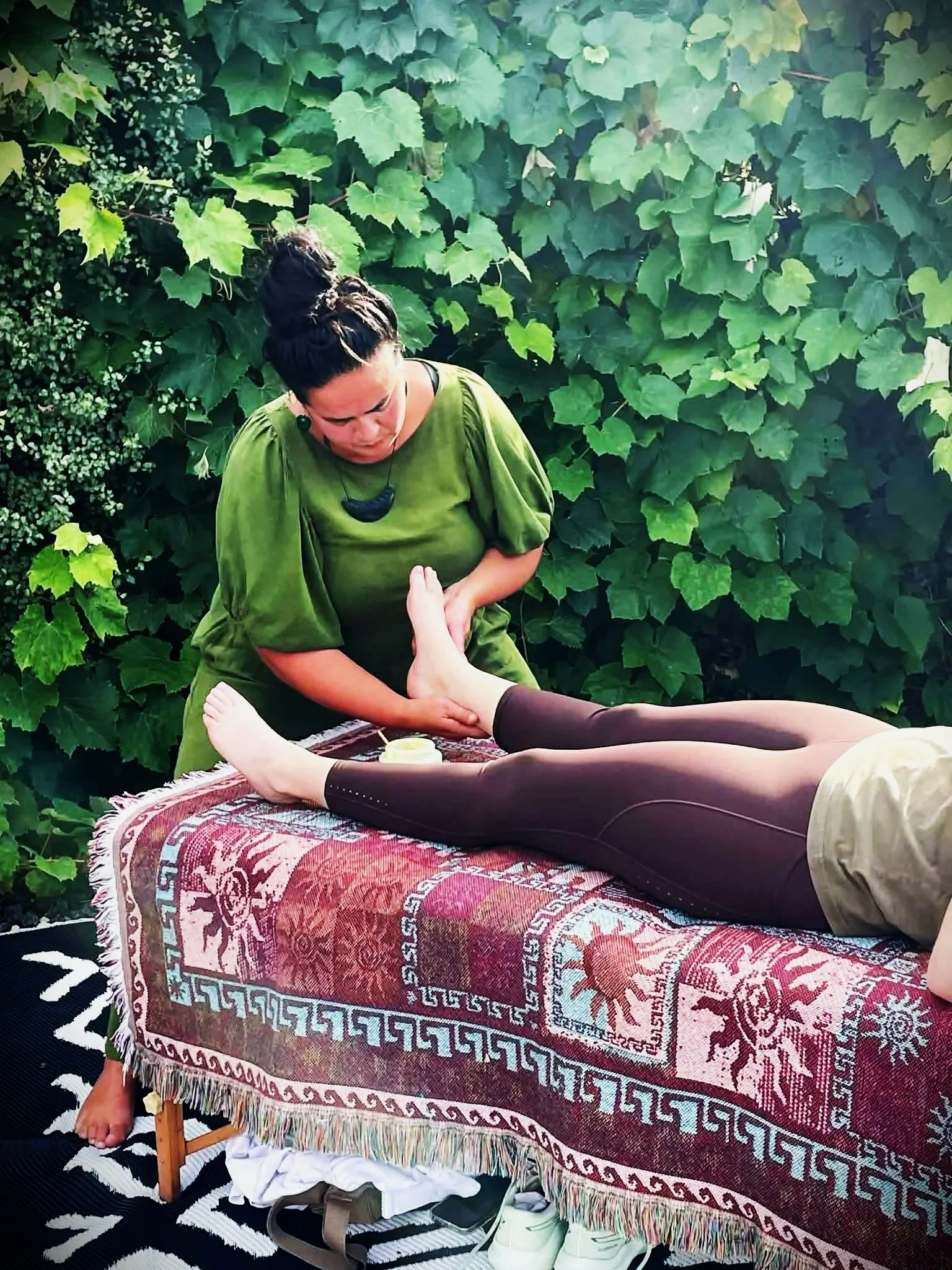 A woman giving a foot massage outdoors on a table with a colorful rug, surrounded by lush green foliage.