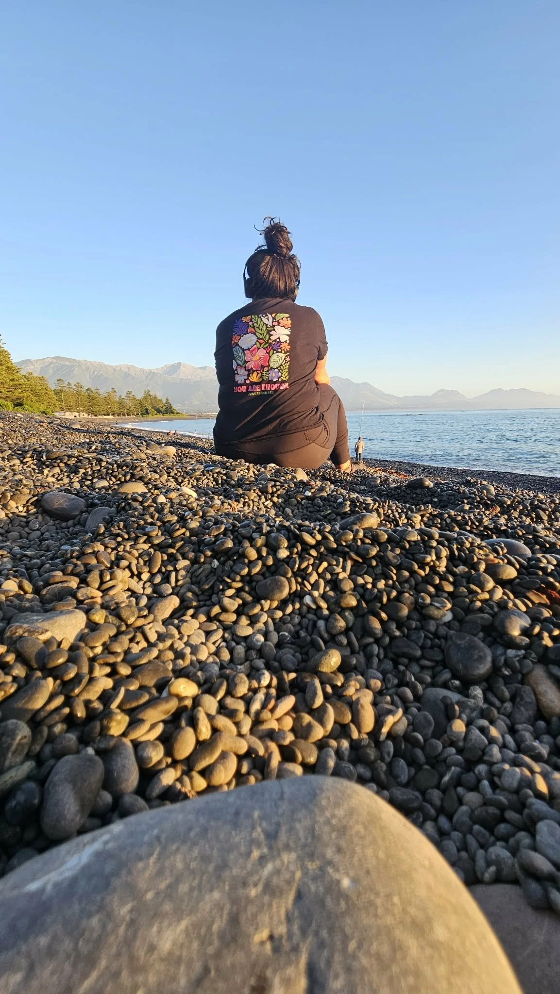 Person sitting on a pebble-covered beach, facing the water with mountains in the distance, during sunset.