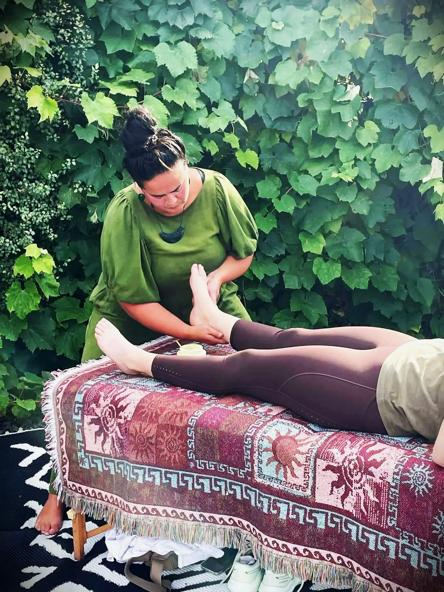 A woman receiving a foot massage outdoors on a table covered with a colorful woven textile, with a backdrop of lush green ivy. The woman is lying on her back, and the masseuse, wearing a green dress, is massaging her feet.