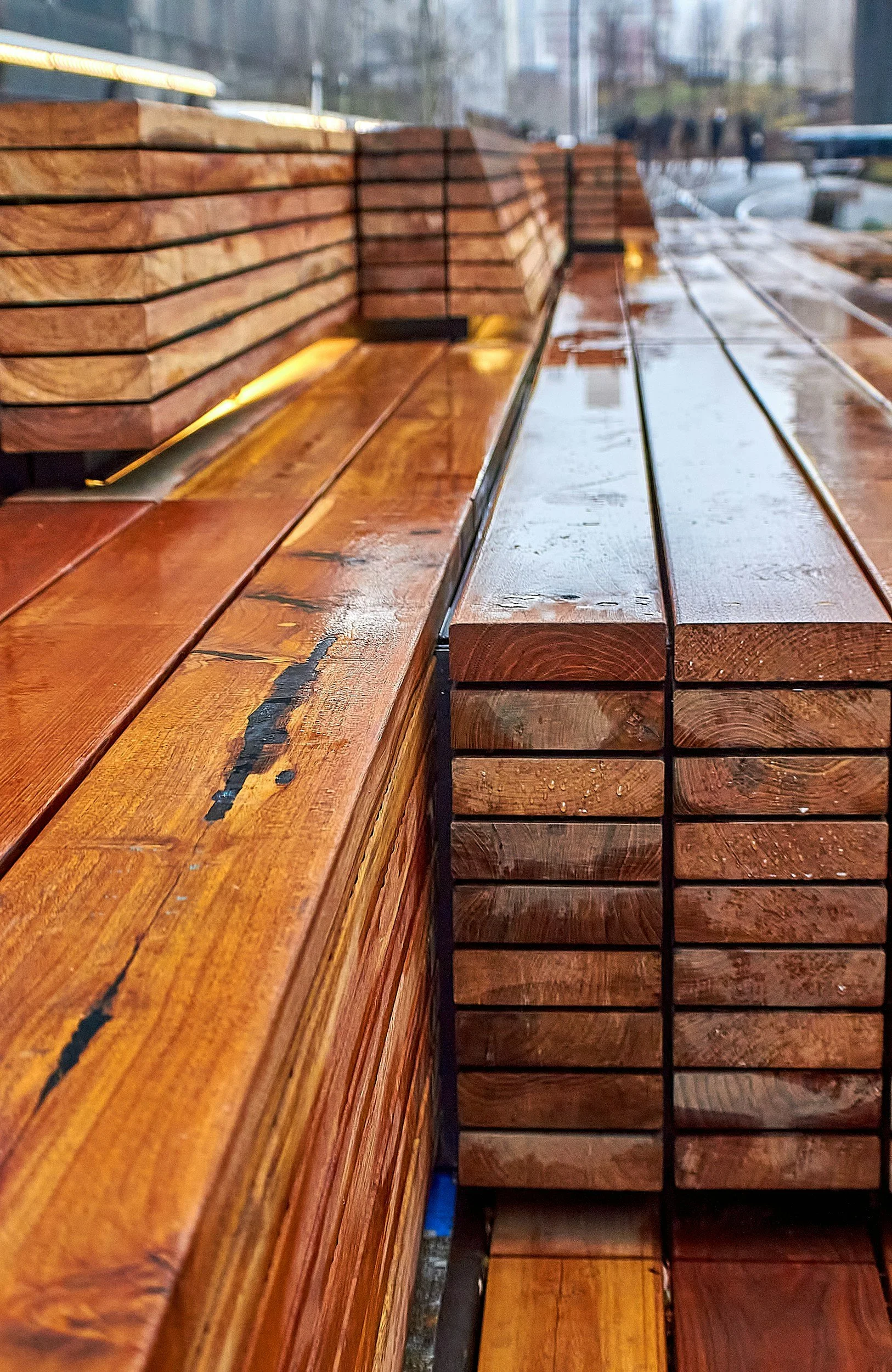 Stacked wooden planks and long wooden boards, some with visible grain and knots, outdoors on a rainy day.