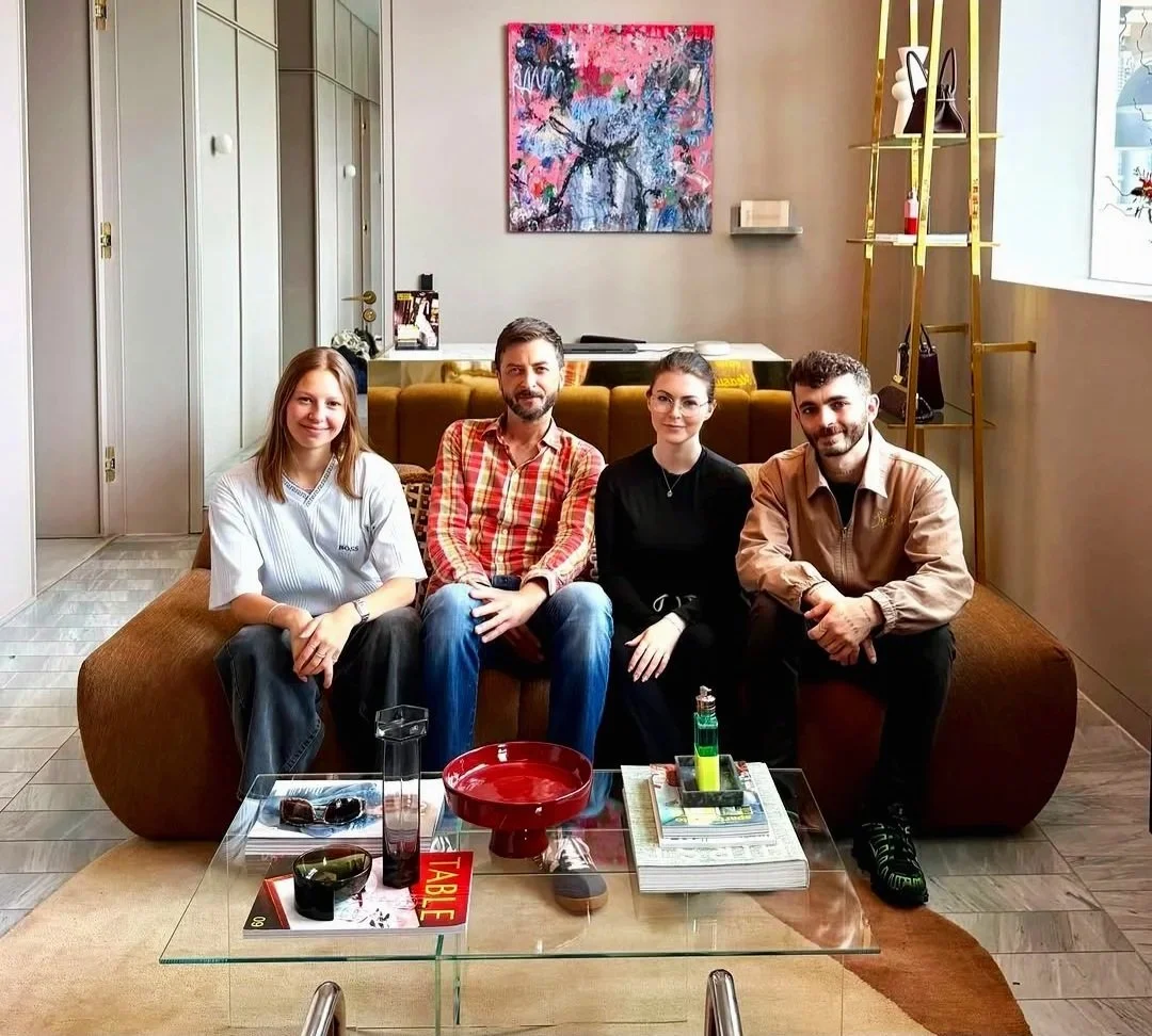 Four people sitting on a brown couch in a contemporary living room, smiling at the camera. There is a glass table in front of them with decorative items, a red bowl, and books. Background includes a colorful abstract painting, a yellow shelving unit with decor and bags, and a window with natural light.