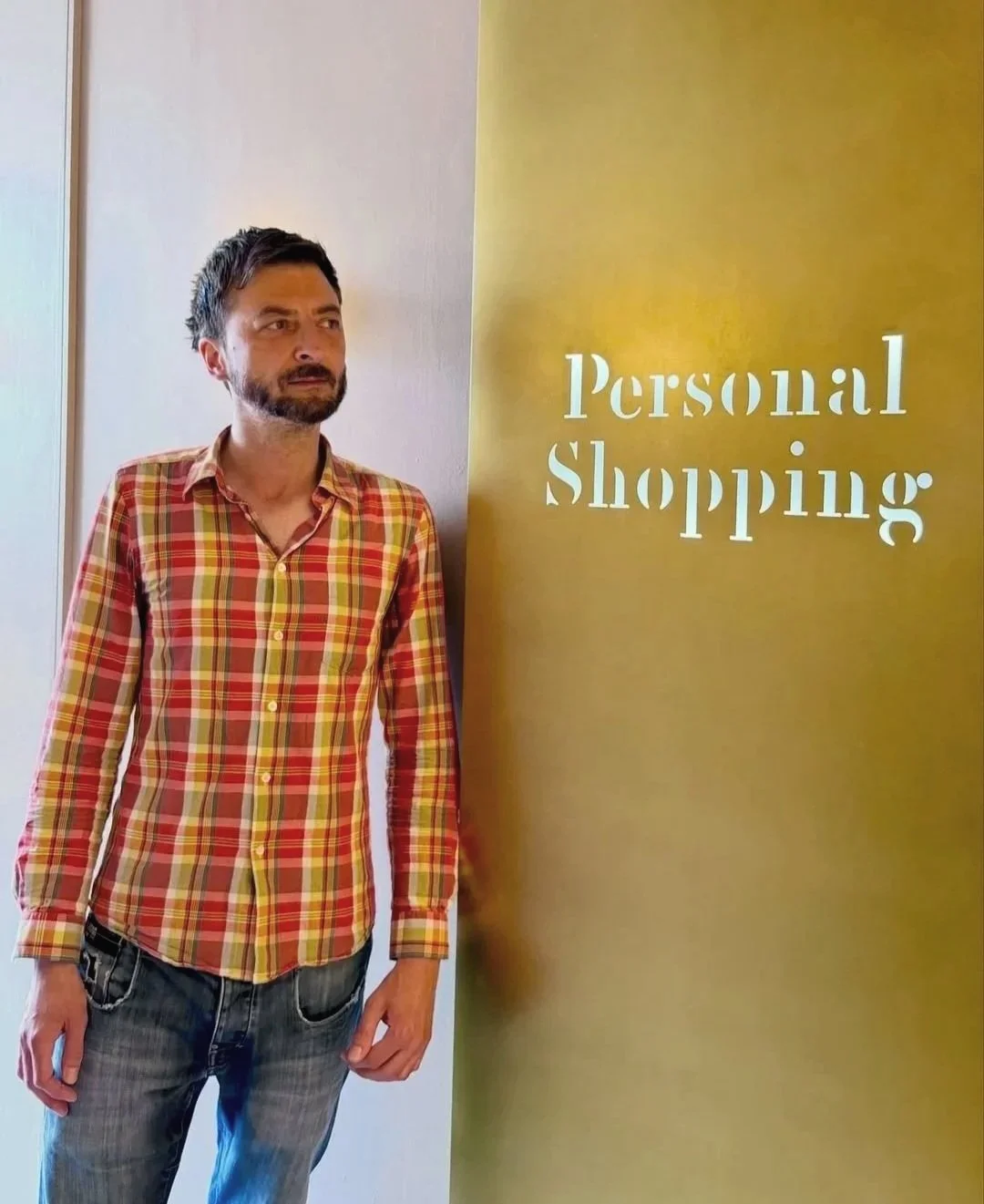 A man with dark hair and a beard wearing a red, yellow, and green plaid shirt and blue jeans standing next to a yellow wall with the words 'Personal Shopping' written on it.