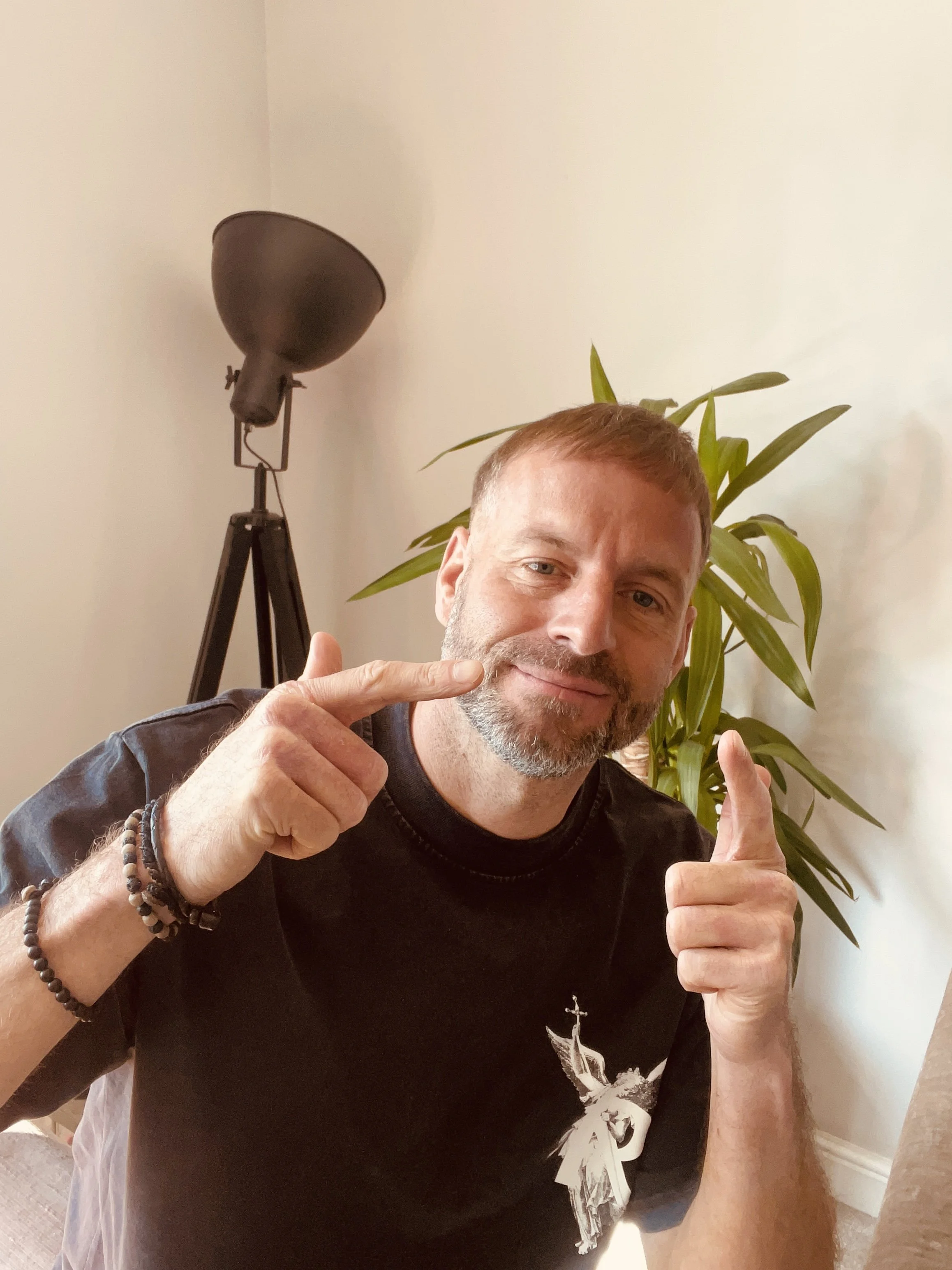 Smiling man with short reddish hair and beard pointing to his cheek and finger. He is wearing a black t-shirt and sits in front of a white wall and a tall green plant, with a large, black, adjustable floor lamp in the background.