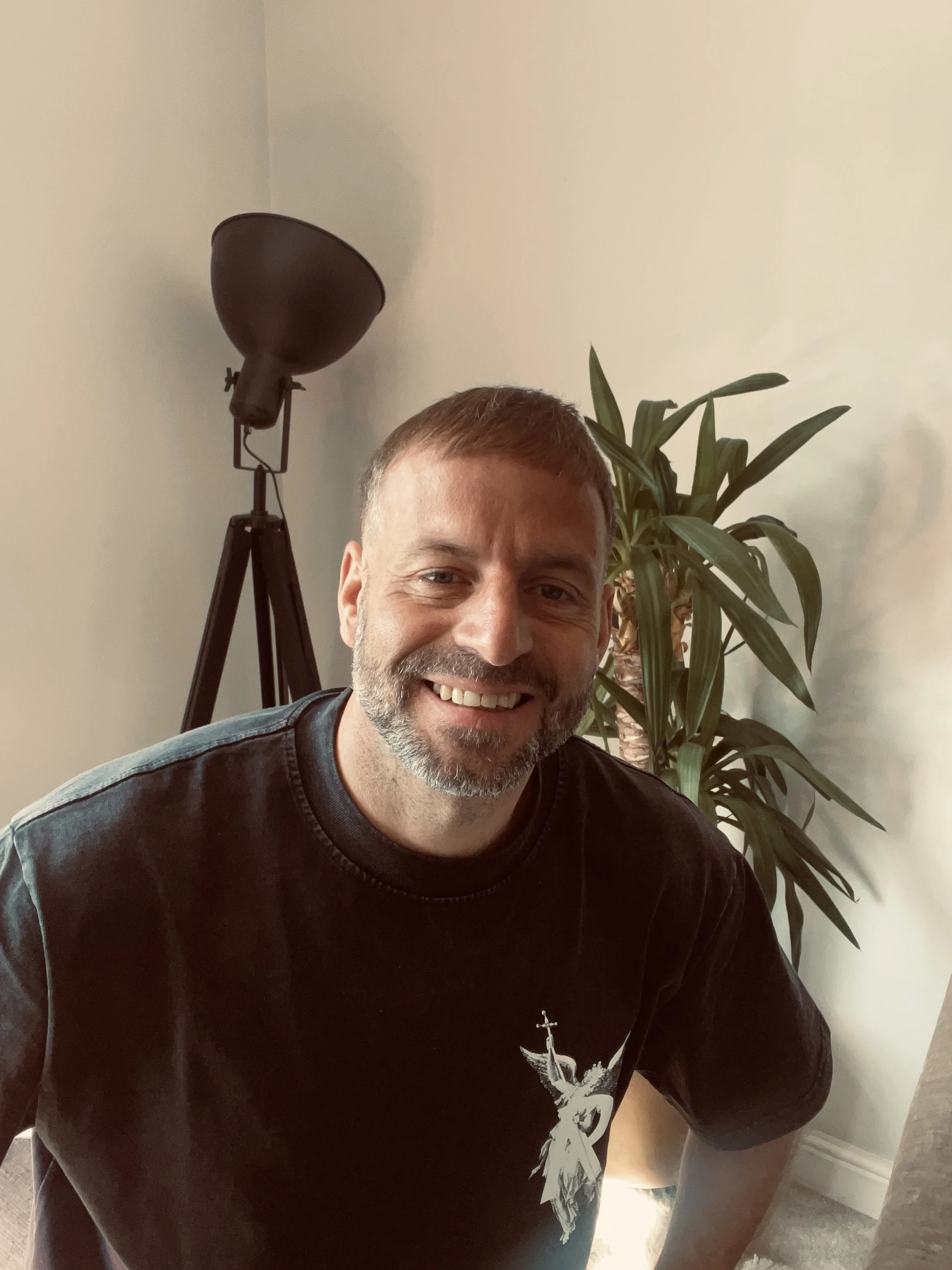 A smiling man with short brown hair, a beard, wearing a black T-shirt with a graphic on it, sitting in a room with a large green plant and a black floor lamp in the background.