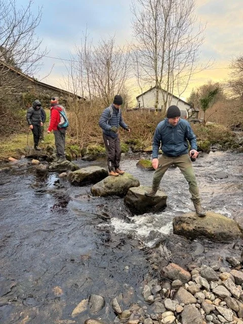 Four people crossing a shallow creek on large rocks during daytime.