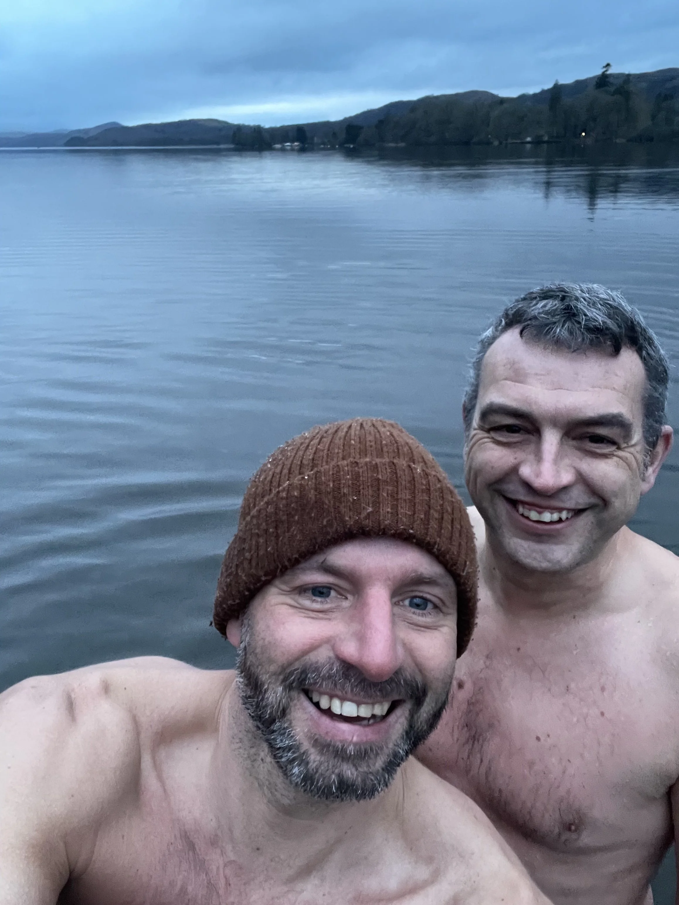Two shirtless men smiling while taking a selfie in a lake with mountains in the background.
