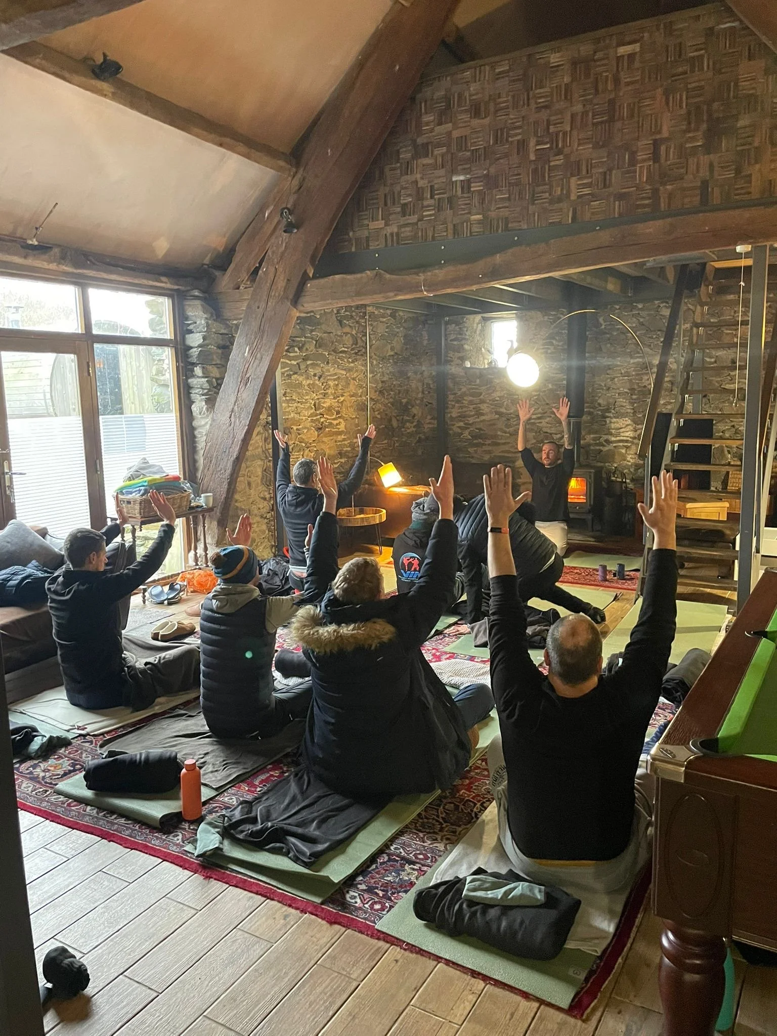 Group of people participating in a yoga or meditation class in a cozy rustic room, with participants seated on mats and raising their hands toward the instructor at the front.
