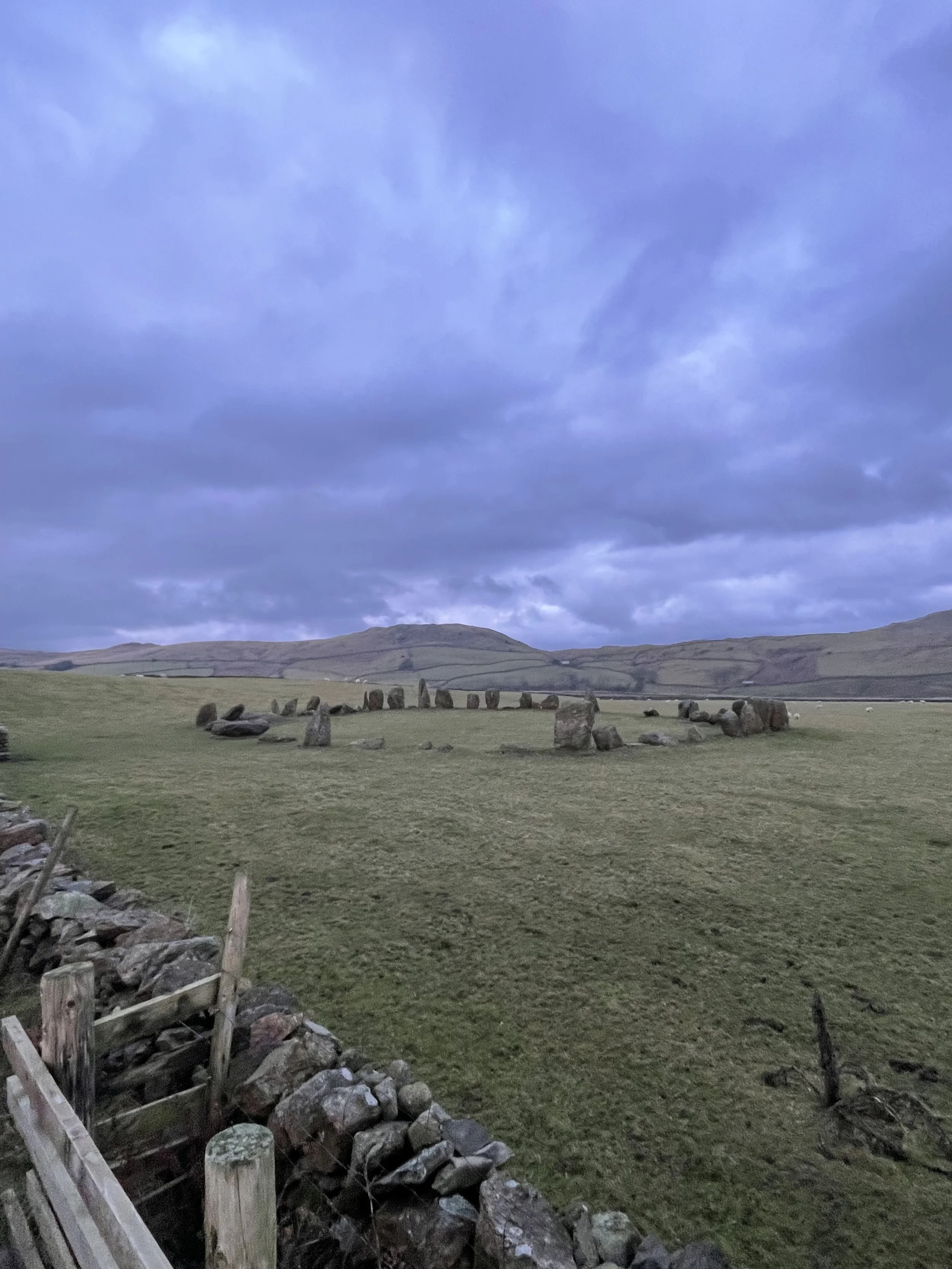 A wide view of a grassy field with prehistoric standing stones arranged in a circle, under a cloudy sky with rolling hills in the background.