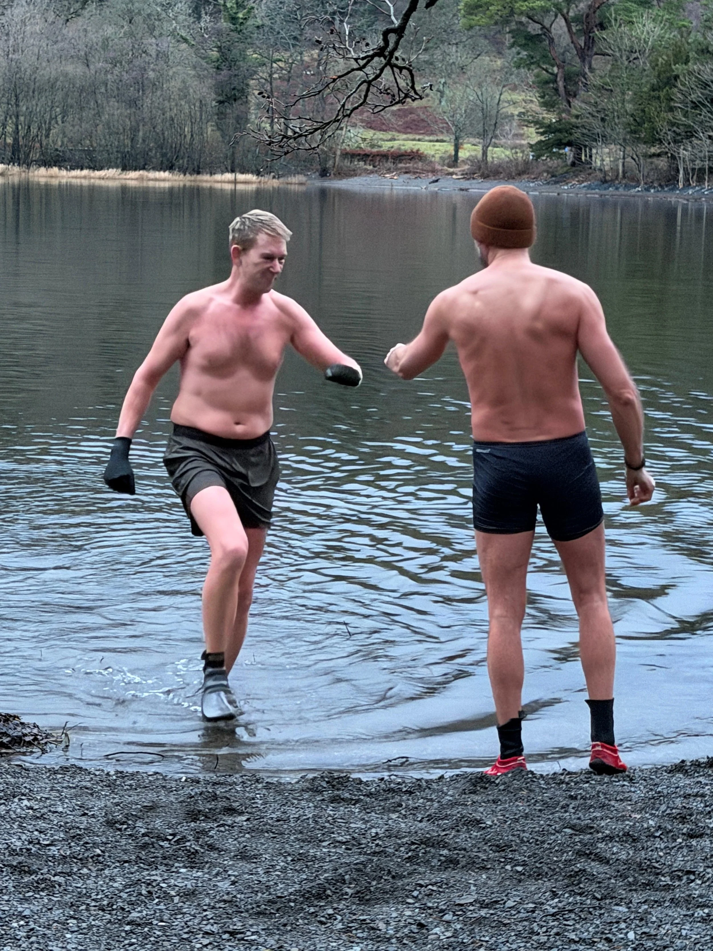 Two shirtless men, one wearing a brown beanie and the other with gloves, are at the edge of a lake engaging in an activity that involves a fist bump or handshake, with trees and a hillside in the background.