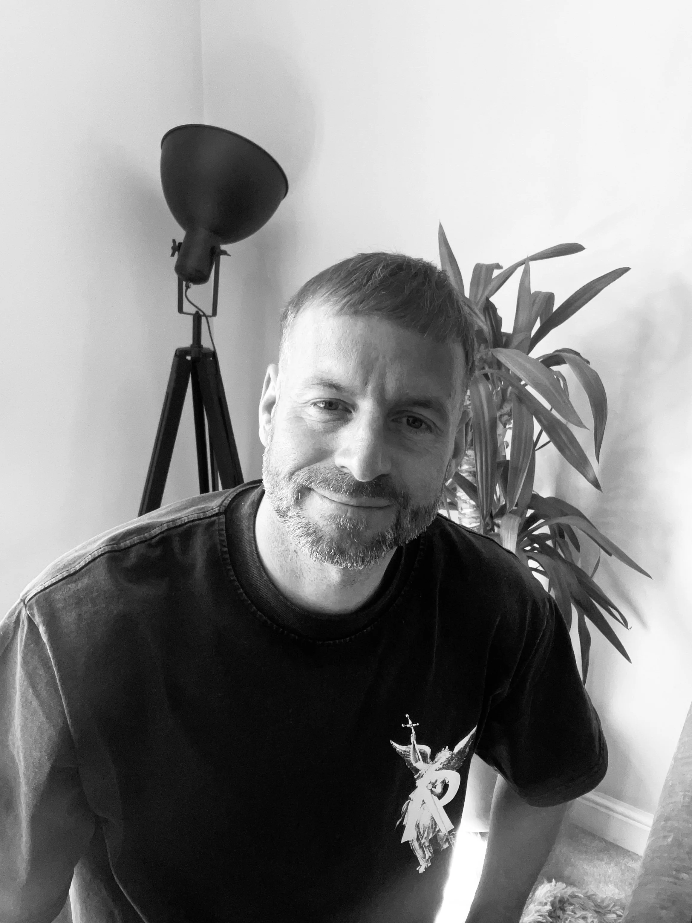 A man with short hair and a beard smiling at the camera, sitting indoors with a tall plant and a floor lamp in the background, in black and white.