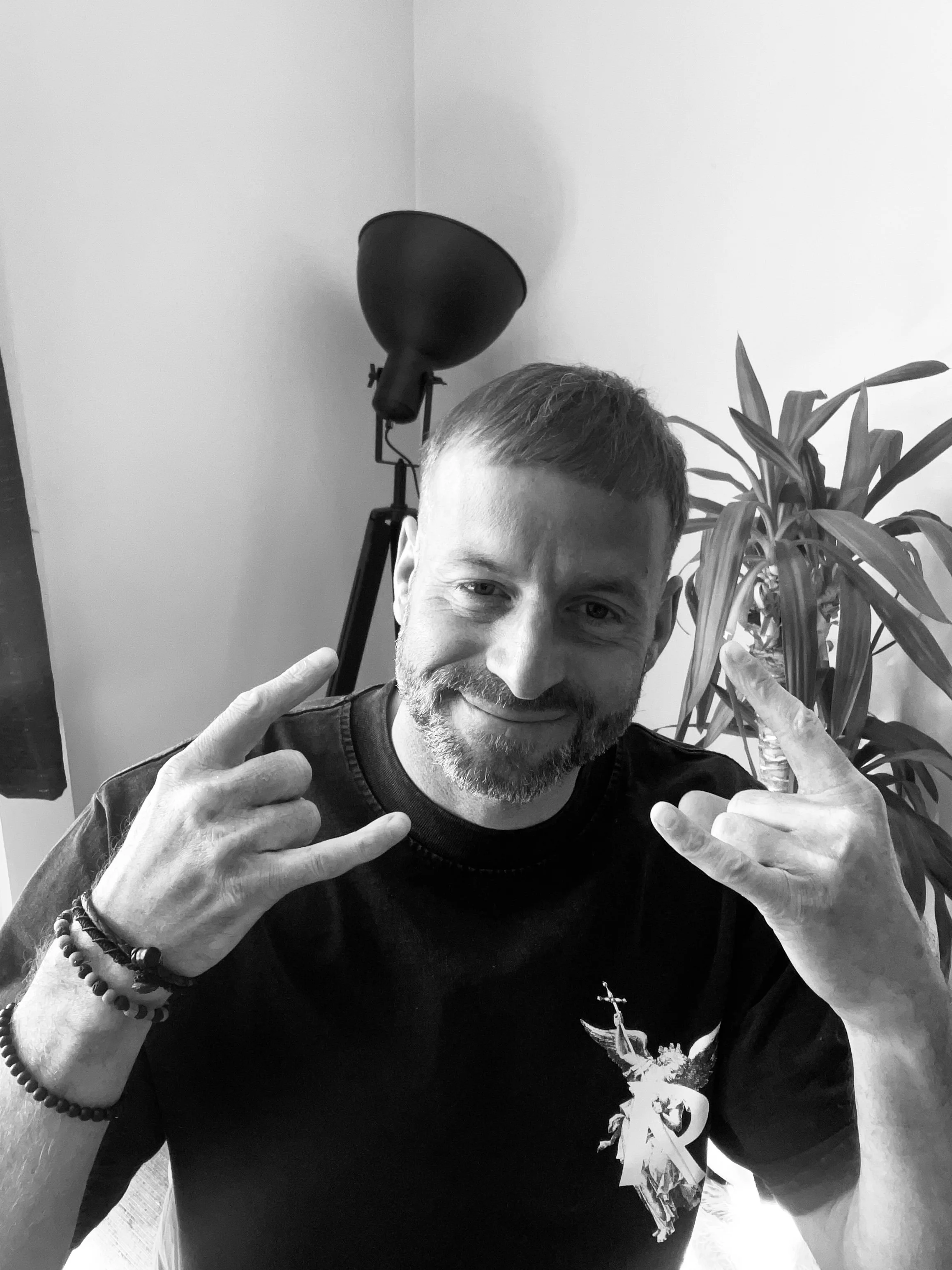 A man with a beard and short hair smiling, making a rock and roll hand gesture with both hands, wearing a dark t-shirt with a graphic design, in a room with a plant and a lamp in the background.