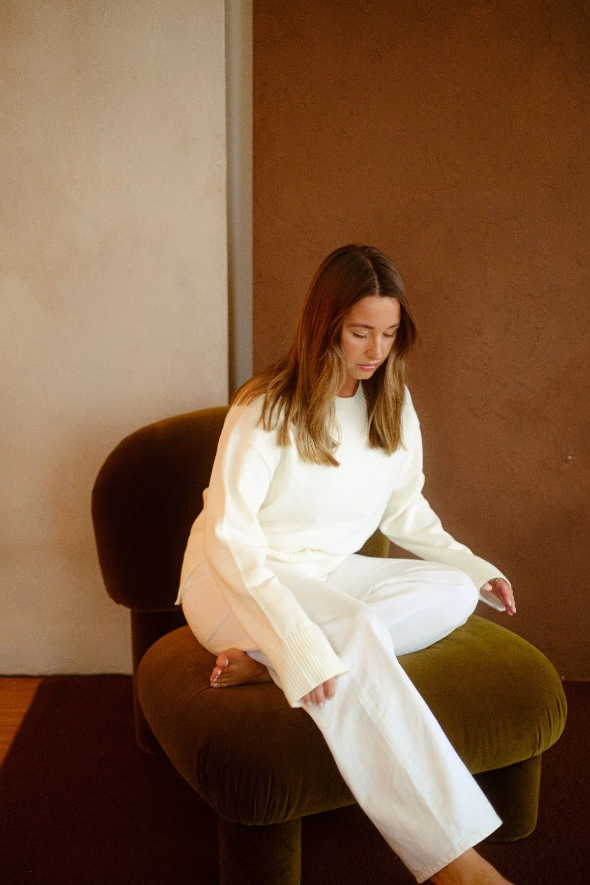 A woman with medium-length brown hair sitting cross-legged on a plush, dark green armchair. She is wearing an oversized white sweater and white pants. She is looking down with a serene expression, her hands resting on her knees. The background features a color-blocked wall with beige and brown tones.