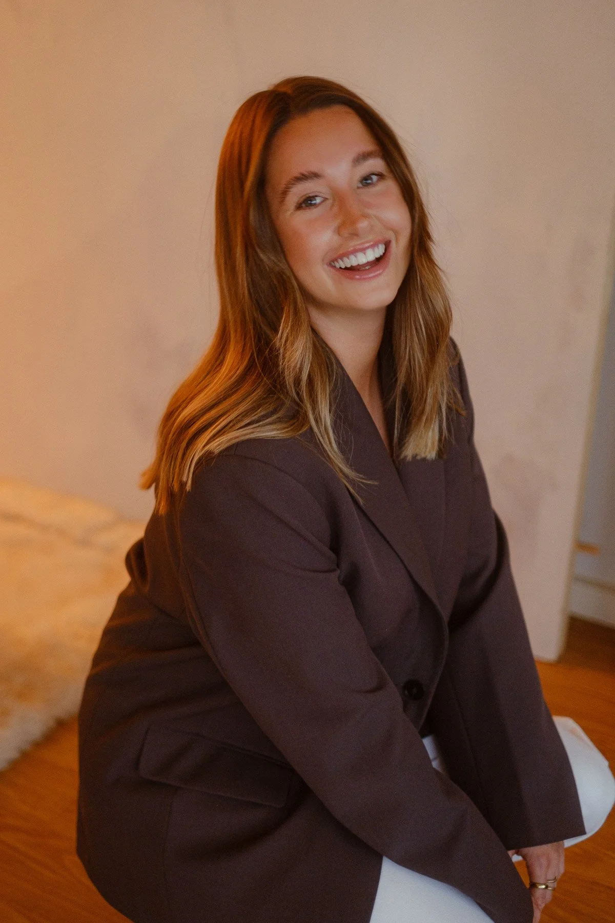 A woman with long blonde hair, wearing a dark blazer, sitting on a chair indoors, smiling at the camera.