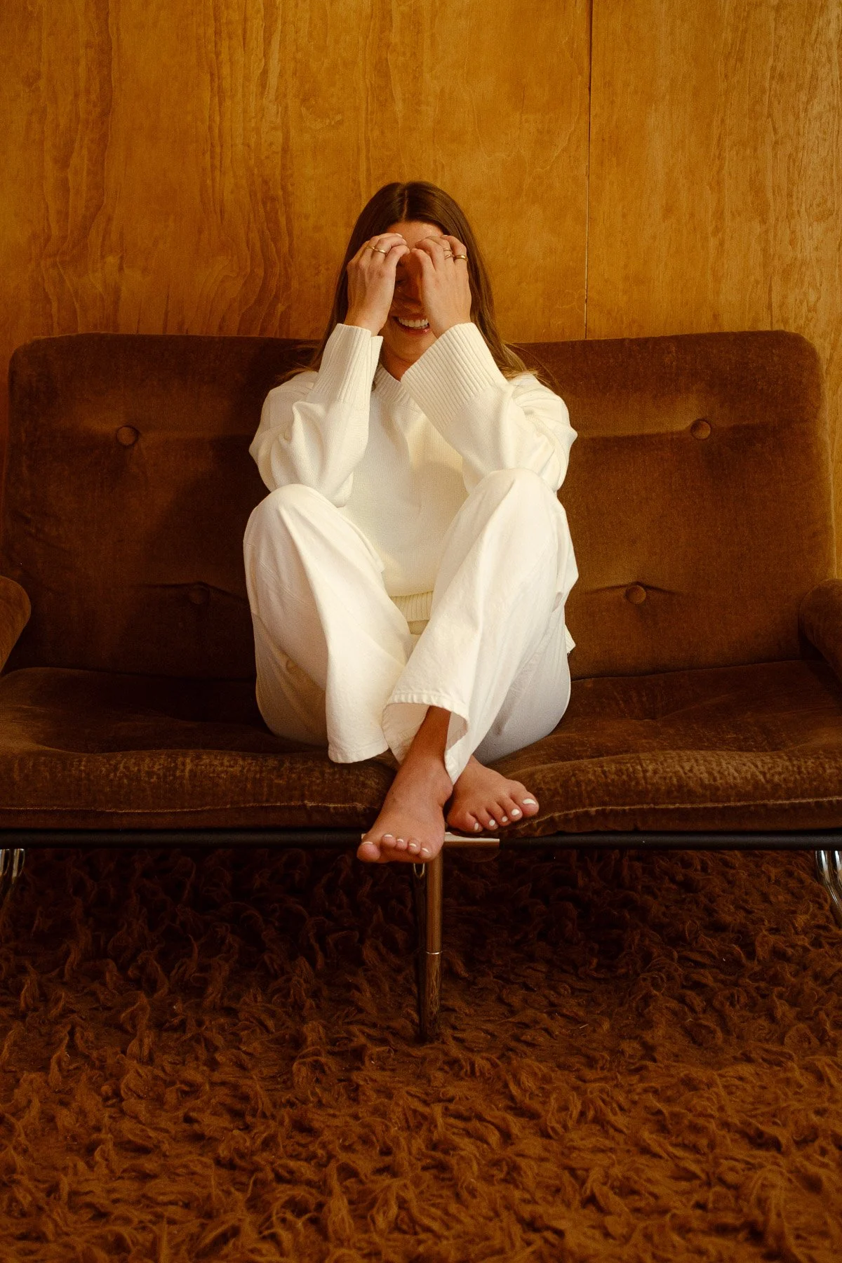A woman sitting on a brown vintage-style sofa with a wooden wall behind her. She is wearing a white sweater and white pants, and is smiling while covering her face with her hands. Her feet are bare on a shaggy brown carpet.