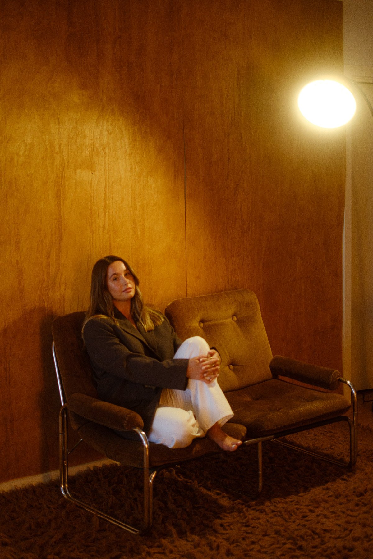 A woman sitting on a vintage brown couch in a room with wooden wall paneling, illuminated by a bright round ceiling light. She has long brown hair, wears a dark blazer, white pants, and barefoot, with her legs crossed and hands resting on her knees.