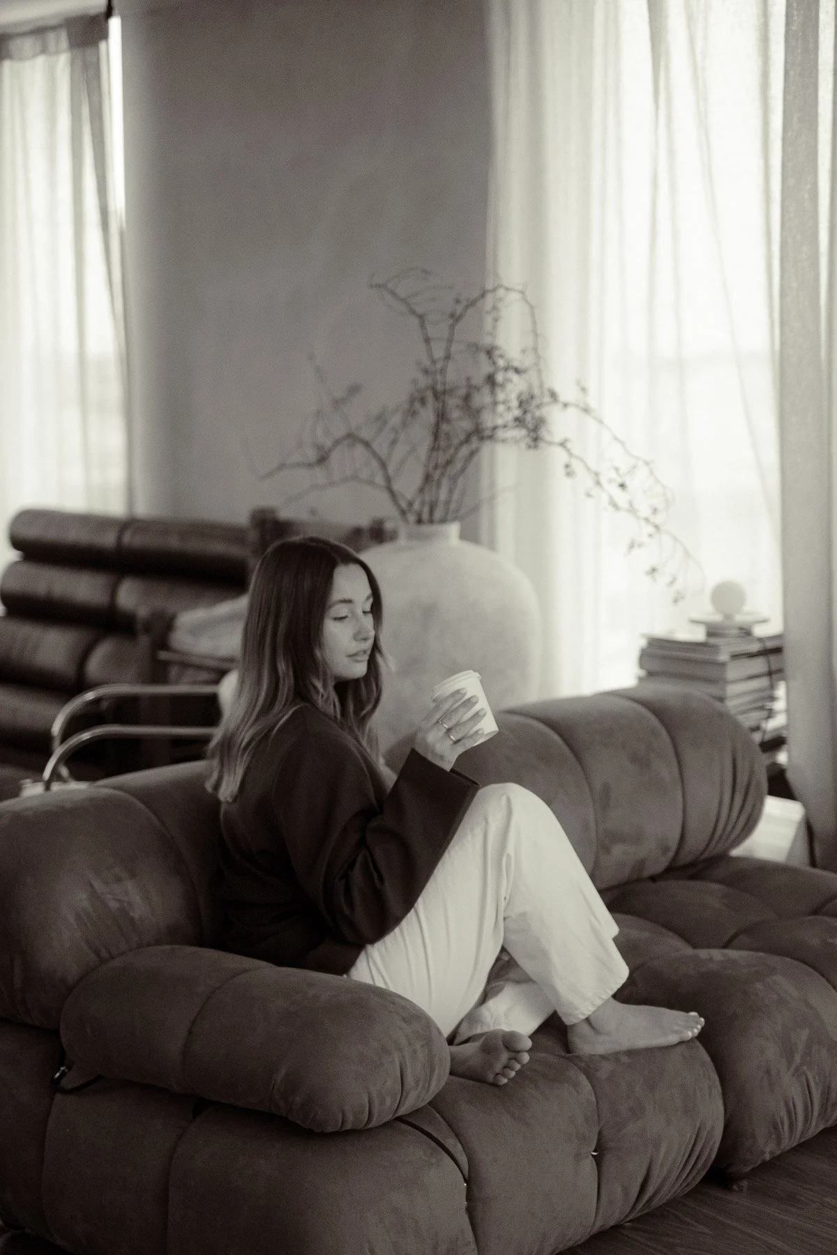 A woman with long hair sits on a plush sofa in a cozy living room, holding a coffee cup and looking down thoughtfully.