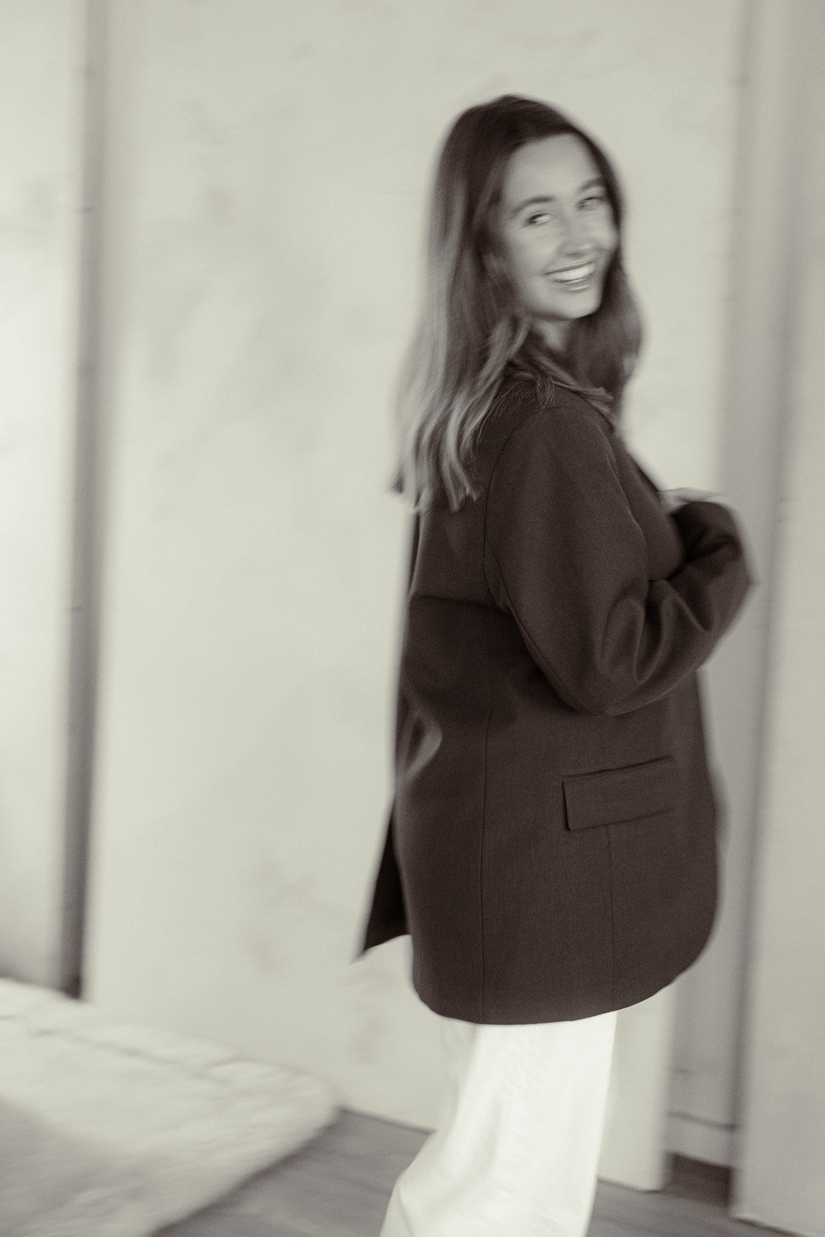 Black and white photo of a woman smiling, wearing a trench coat, standing in front of a plain wall.