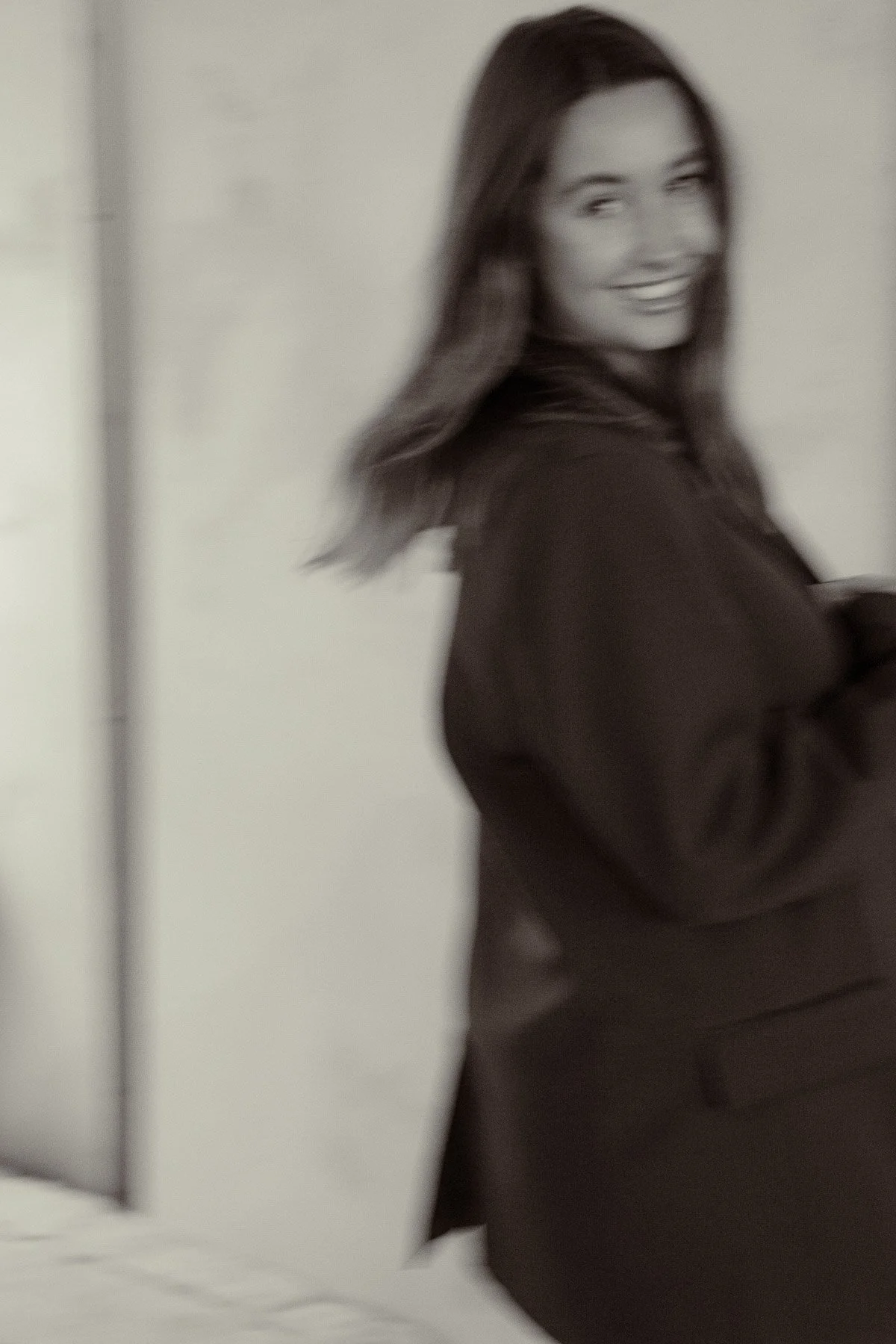 Black and white photo of a smiling woman with long hair, standing inside a room, with her arm slightly bent at her side, wearing a dark long-sleeved top, and standing next to a white wall or door.