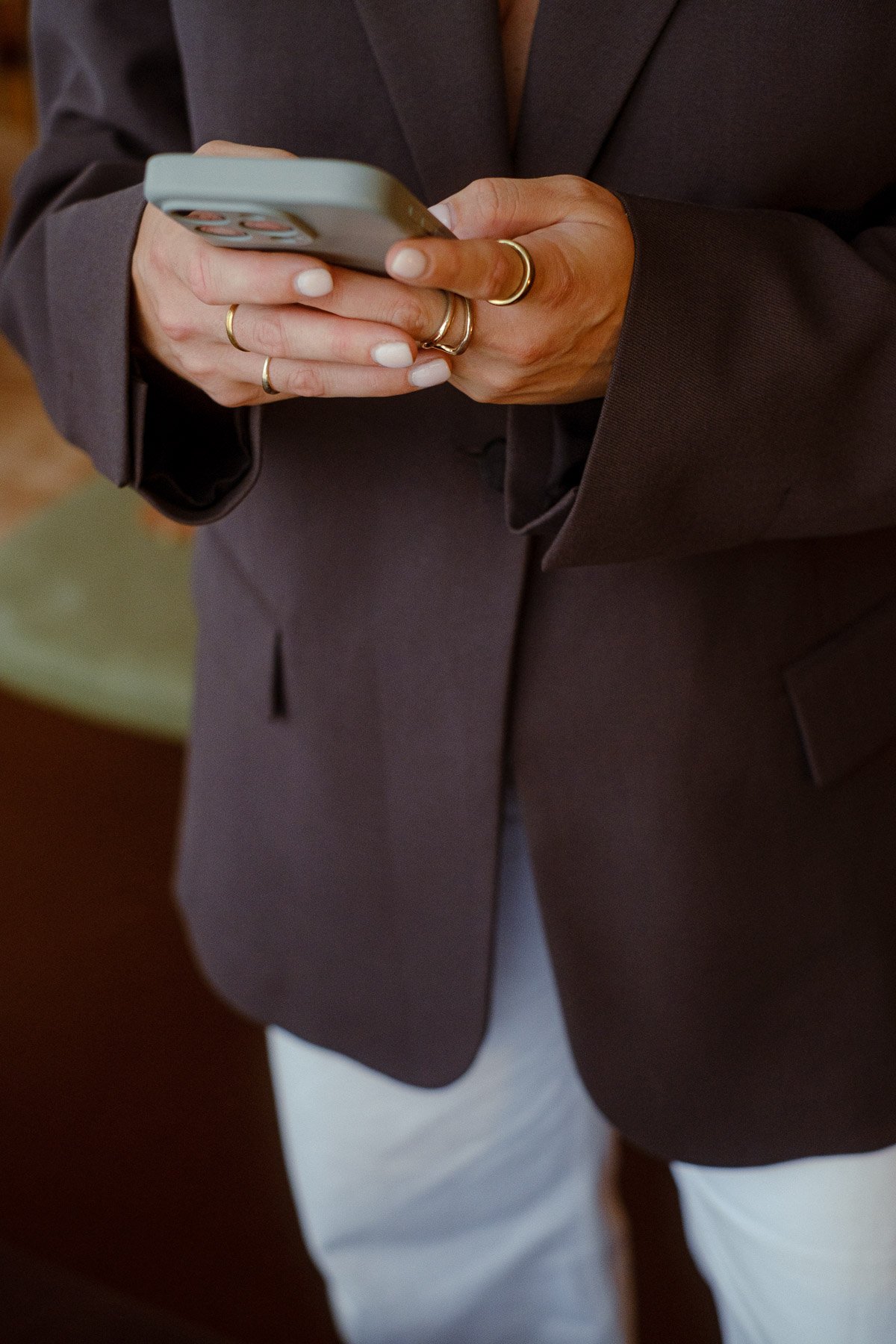 Person wearing a dark blazer and white pants, holding a smartphone in both hands with rings on fingers.