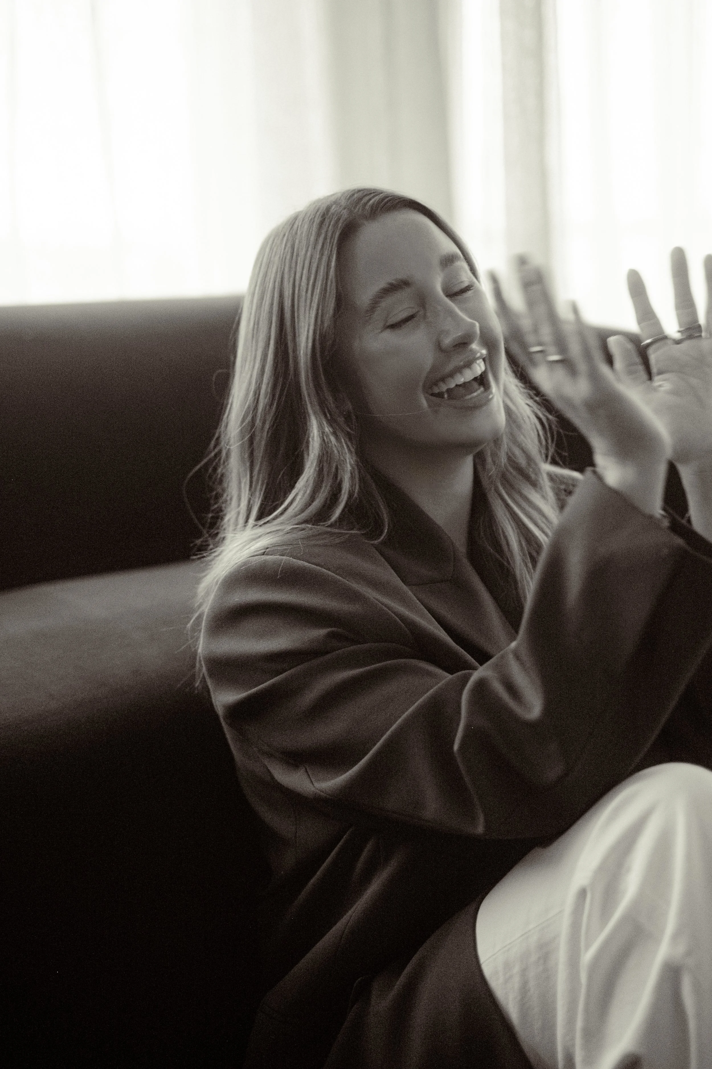 A woman with long hair and rings on her fingers is sitting on a sofa, laughing with her eyes closed, in a room with light curtains.