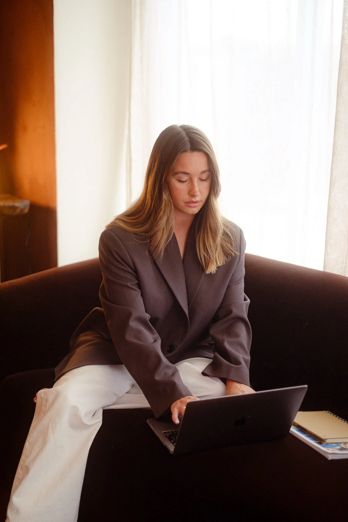 A woman with long light brown hair wearing a brown blazer and cream-colored pants sitting on a black couch, looking at a laptop computer on her lap, in a living room with cream curtains and a wooden shelf.