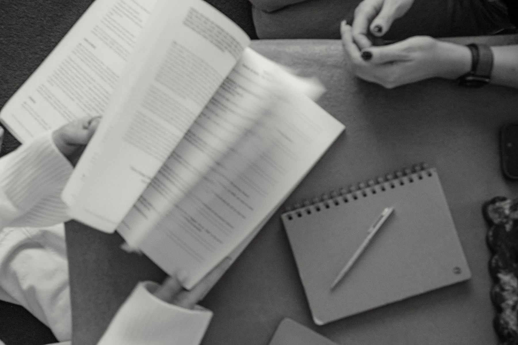 A person holding an open book or notebook in a grayscale photo. The scene includes a desk with a small spiral notebook, a pen, a wristwatch on the person's wrist, and other objects around.