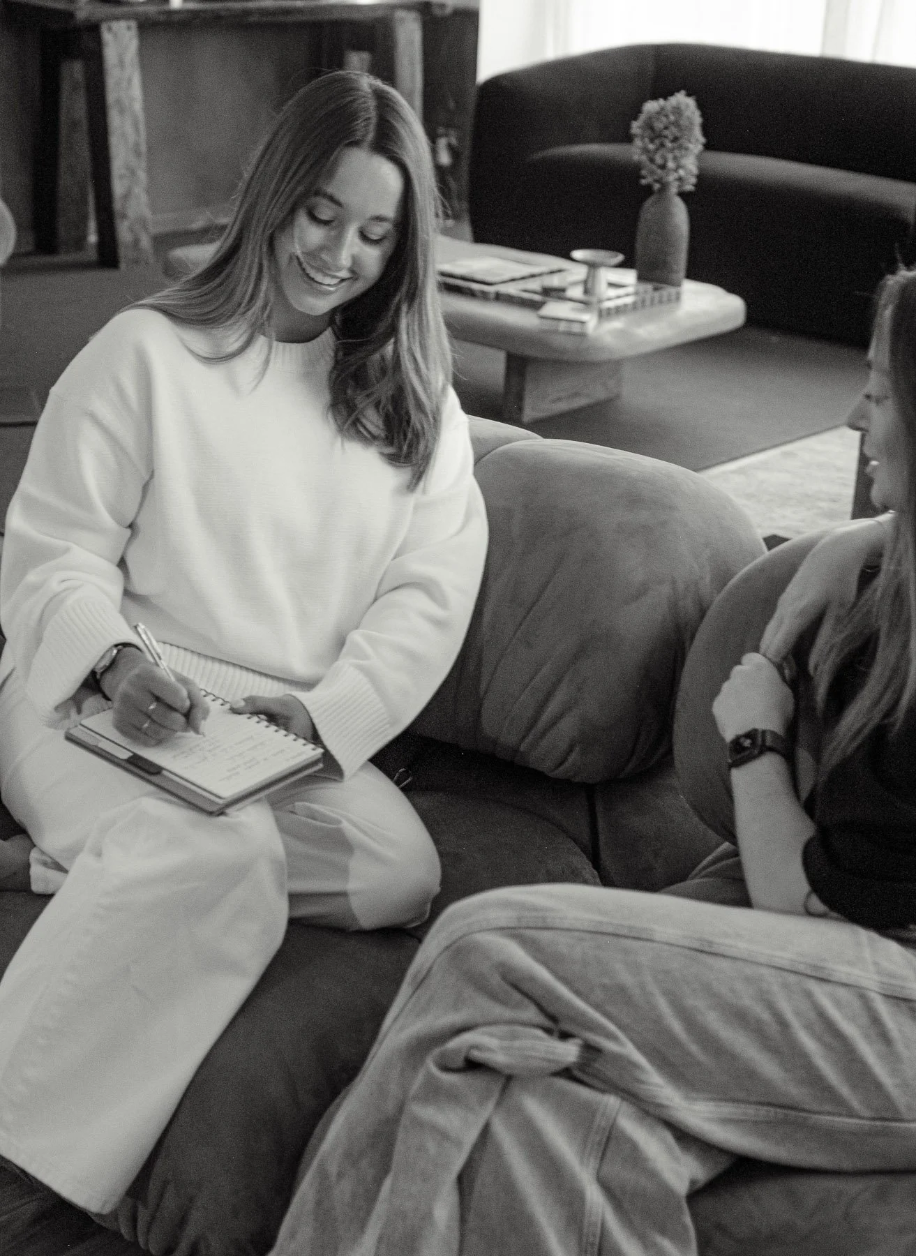 Two women sitting on a sofa having a conversation in a cozy living room. One woman is smiling and writing in a notebook, while the other is listening attentively.