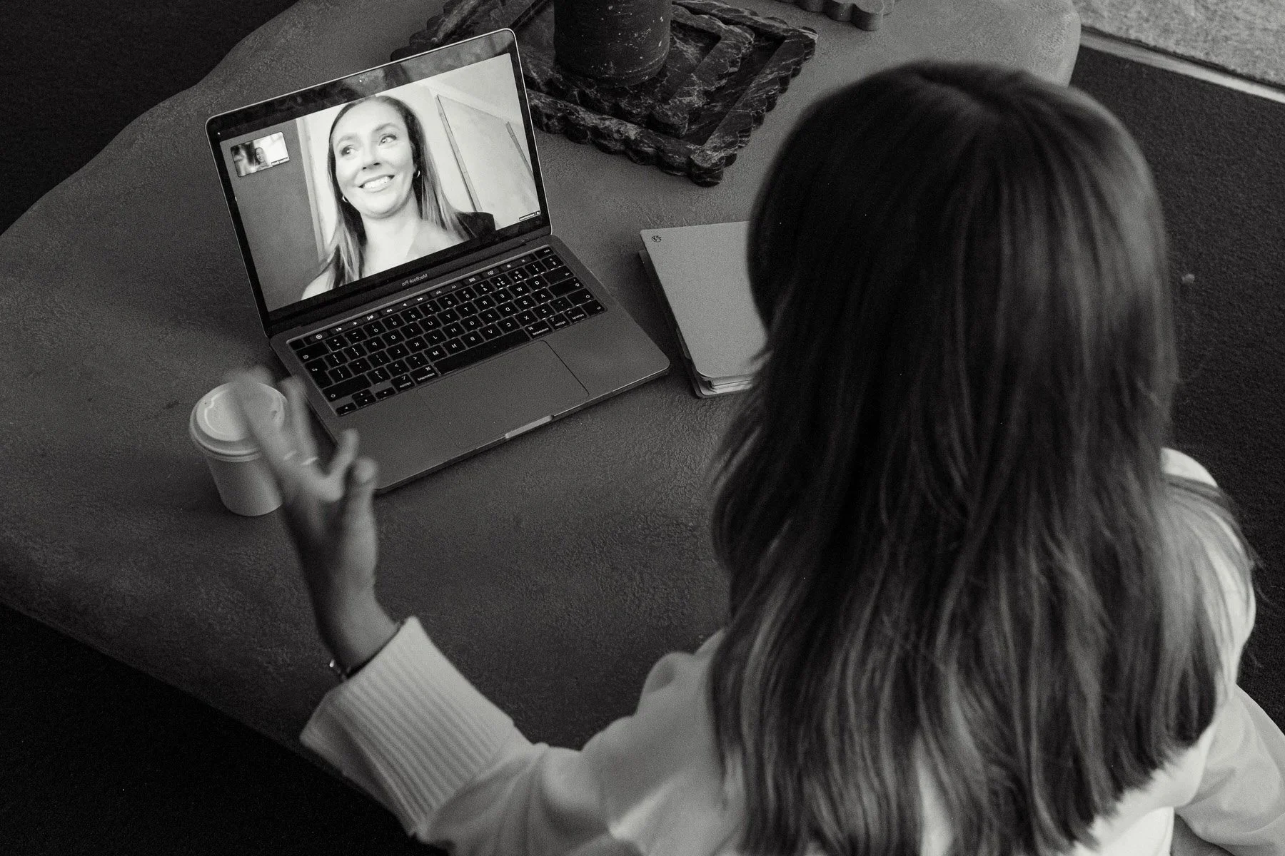 A woman with long hair sitting at a table, participating in a video call on a laptop, smiling at the screen.