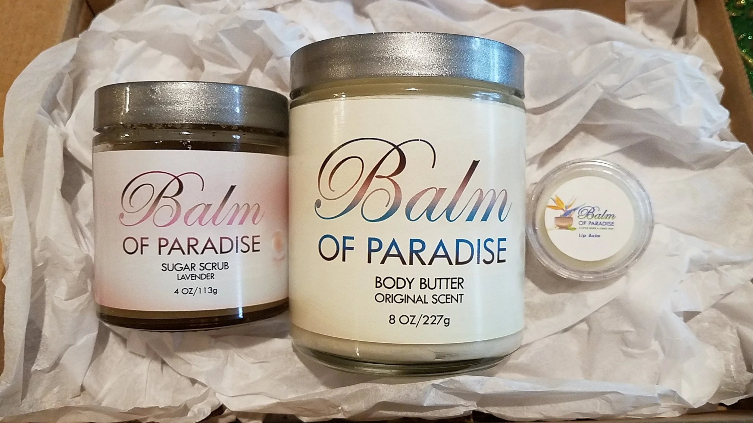 Three cosmetic products from Balm of Paradise placed on crumpled white paper: a lavender sugar scrub, a bottle of body butter with original scent, and a lip balm.