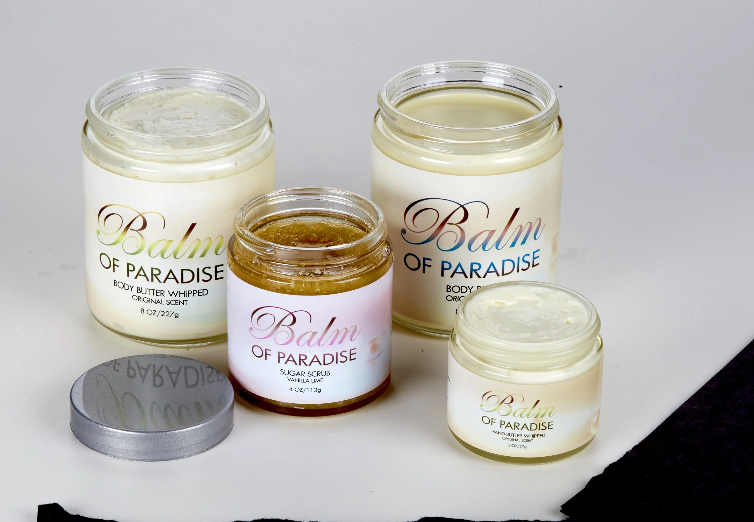 Several jars of Balm of Paradise body butter and sugar scrub, with metal lid and white labels showing brand and scent, placed on a white surface with black paper underneath.