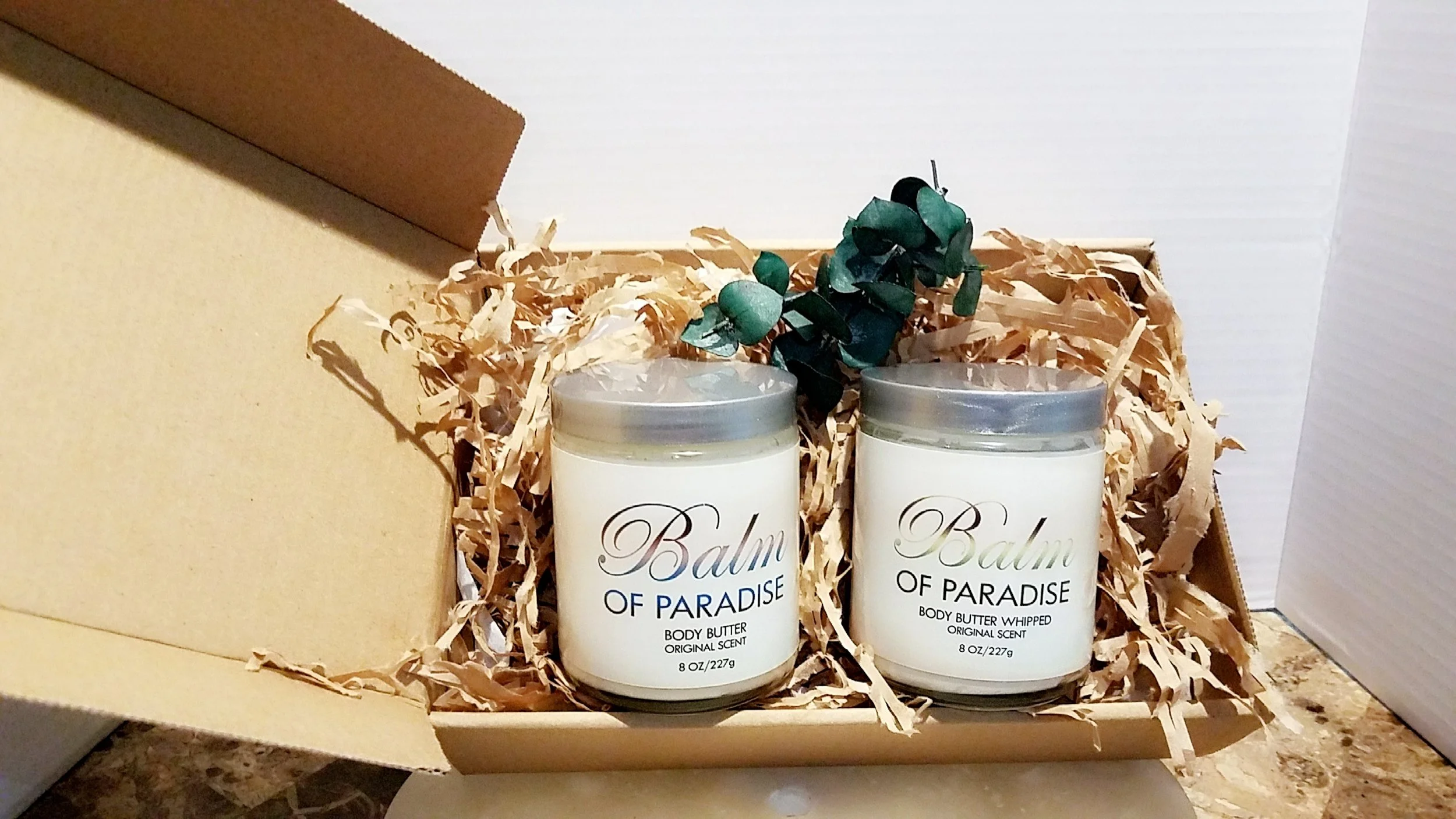 Two white body butter containers labeled 'Balm of Paradise' placed in a cardboard box with shredded paper filling.