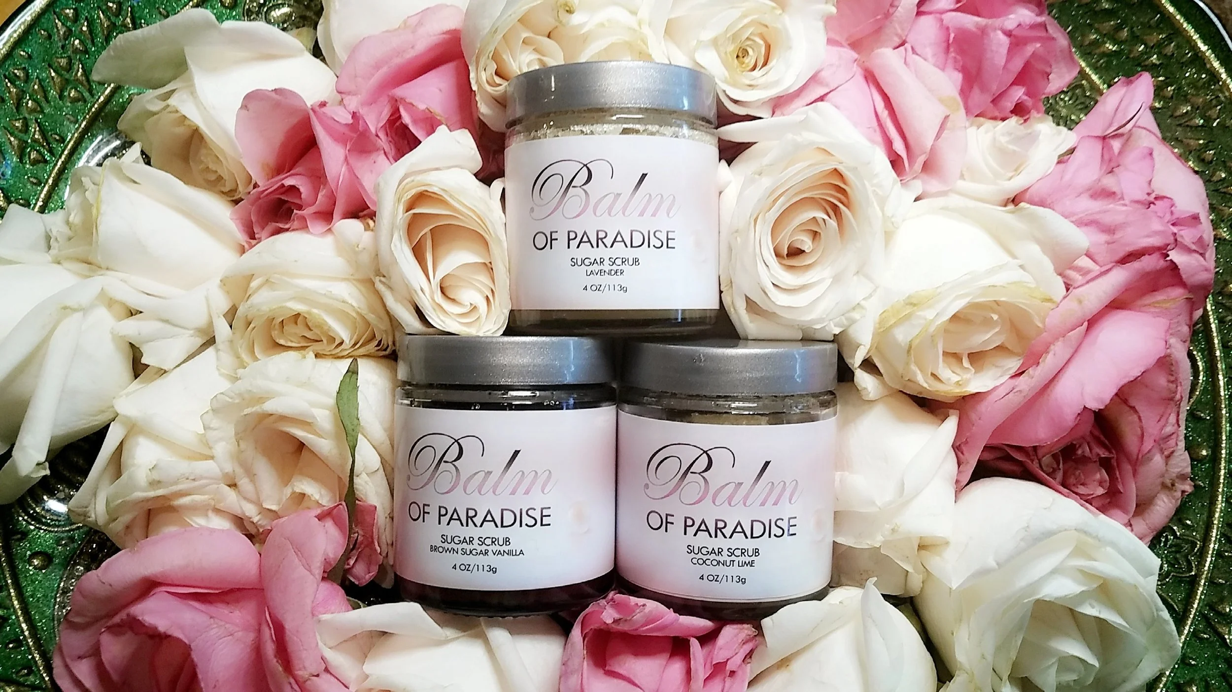 A decorative tray filled with pink and white rose petals, holding several small jars of Balm of Paradise sugar scrub in lavender, vanilla, coconut lime, and brown sugar vanilla scents.