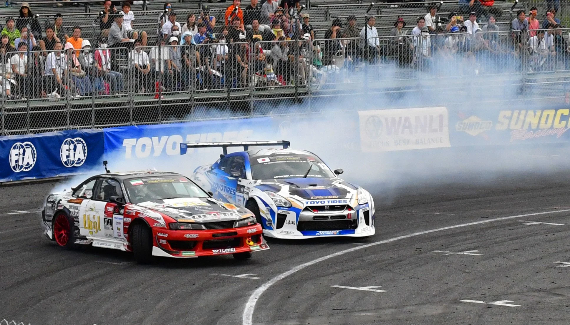 Two race cars drifting on a track with smoke, crowd watching from stands in the background.
