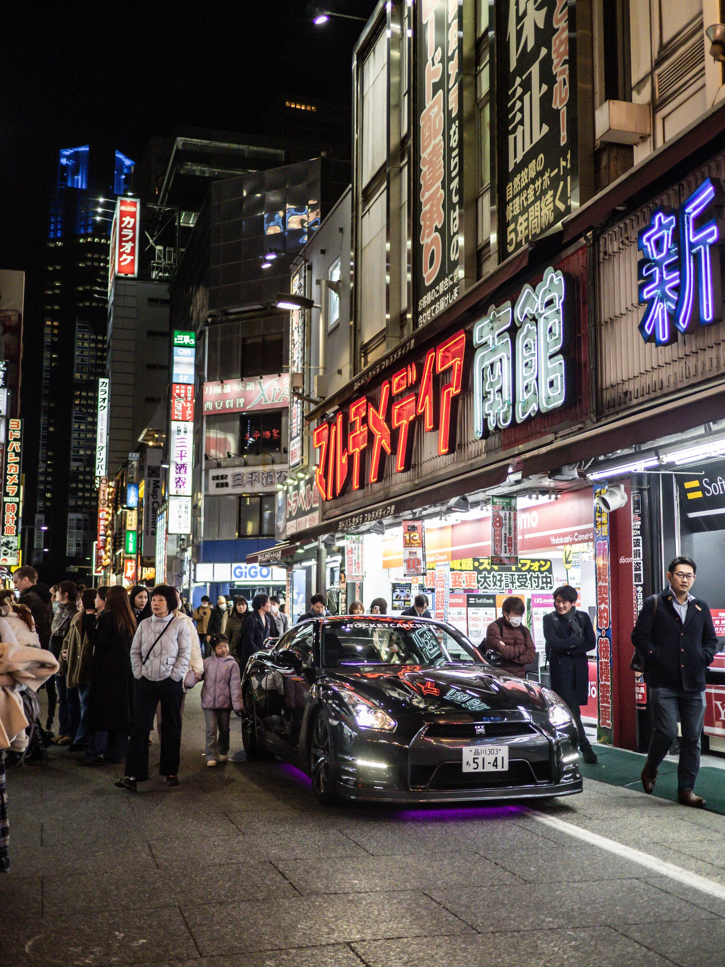 Night scene of busy street in Japan with neon signs, crowds, and a black sports car parked on the side of the street.