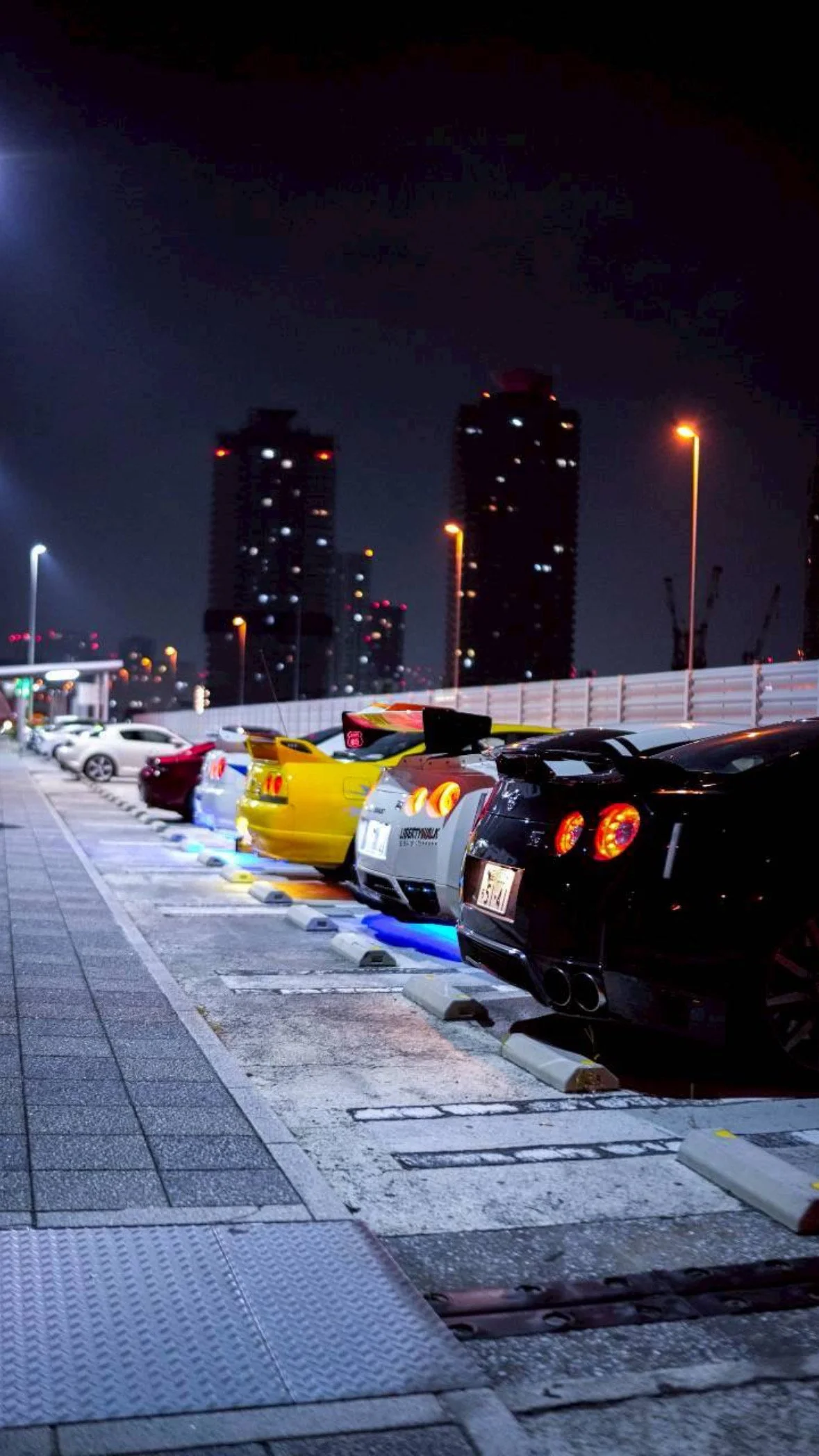 Nighttime view of a parking lot with sports cars, including a yellow car, white car, red car, and black car, illuminated by blue and yellow lights with tall buildings in the background.