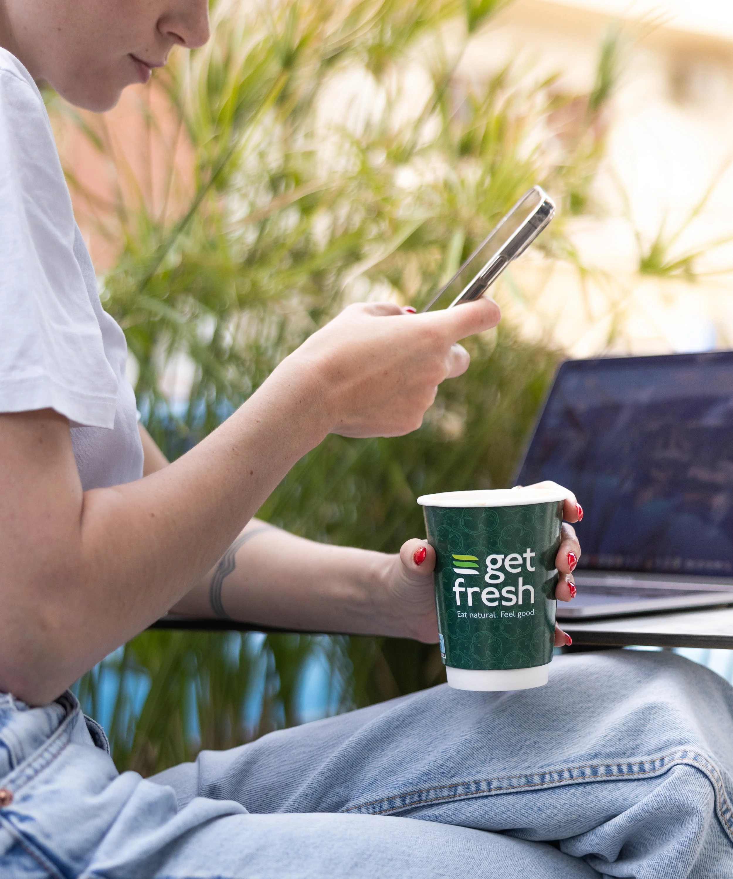 A stylish, cool person in a white shirt and blue jeans sitting down, holding a green cup with the text "get fresh" and an illustration of leaves. The person is looking at their phone, with greenery in the background.