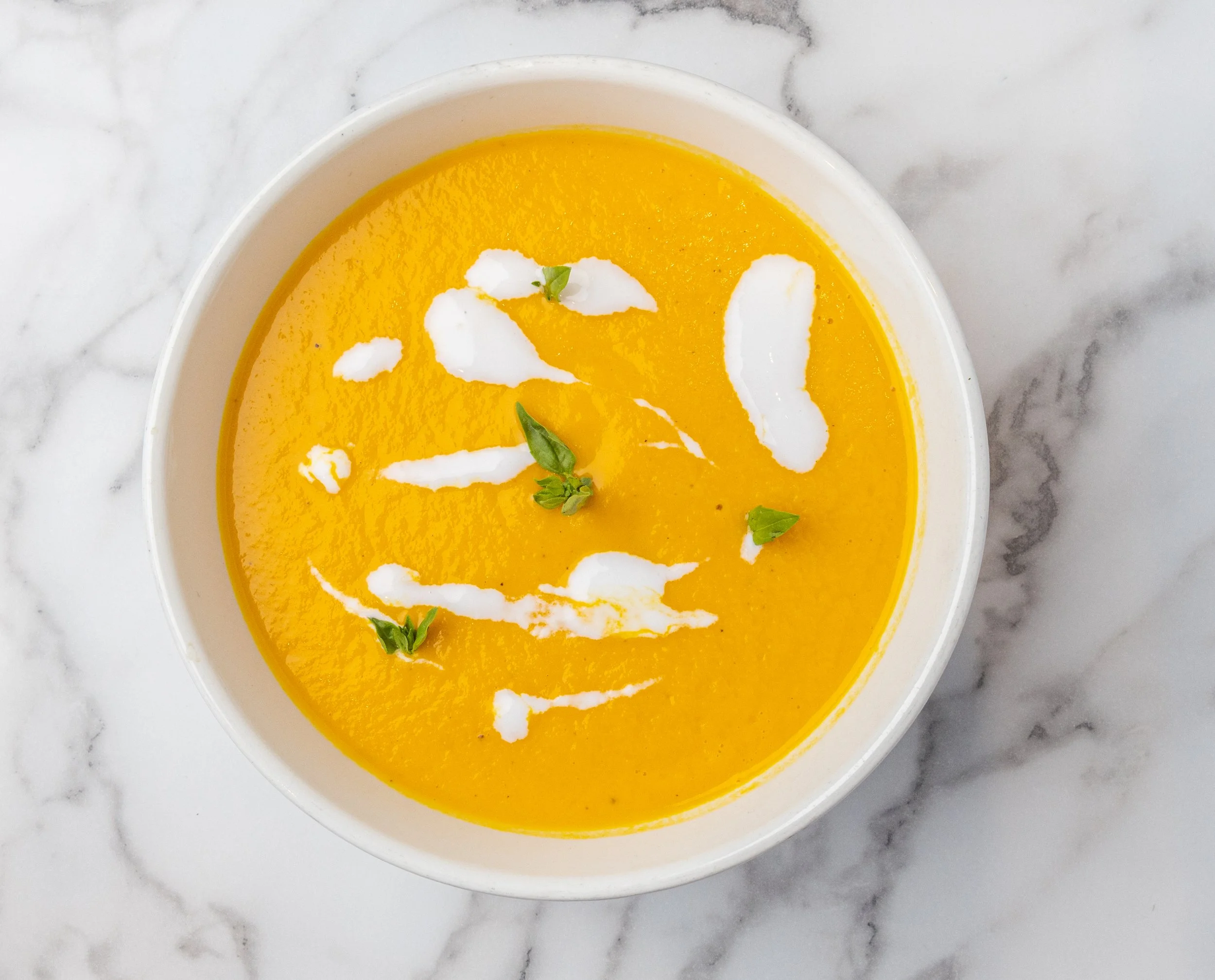 A bowl of bright orange pumpkin soup garnished with swirls of cream and small green herbs, placed on a white marble surface. Looks delicious and healthy