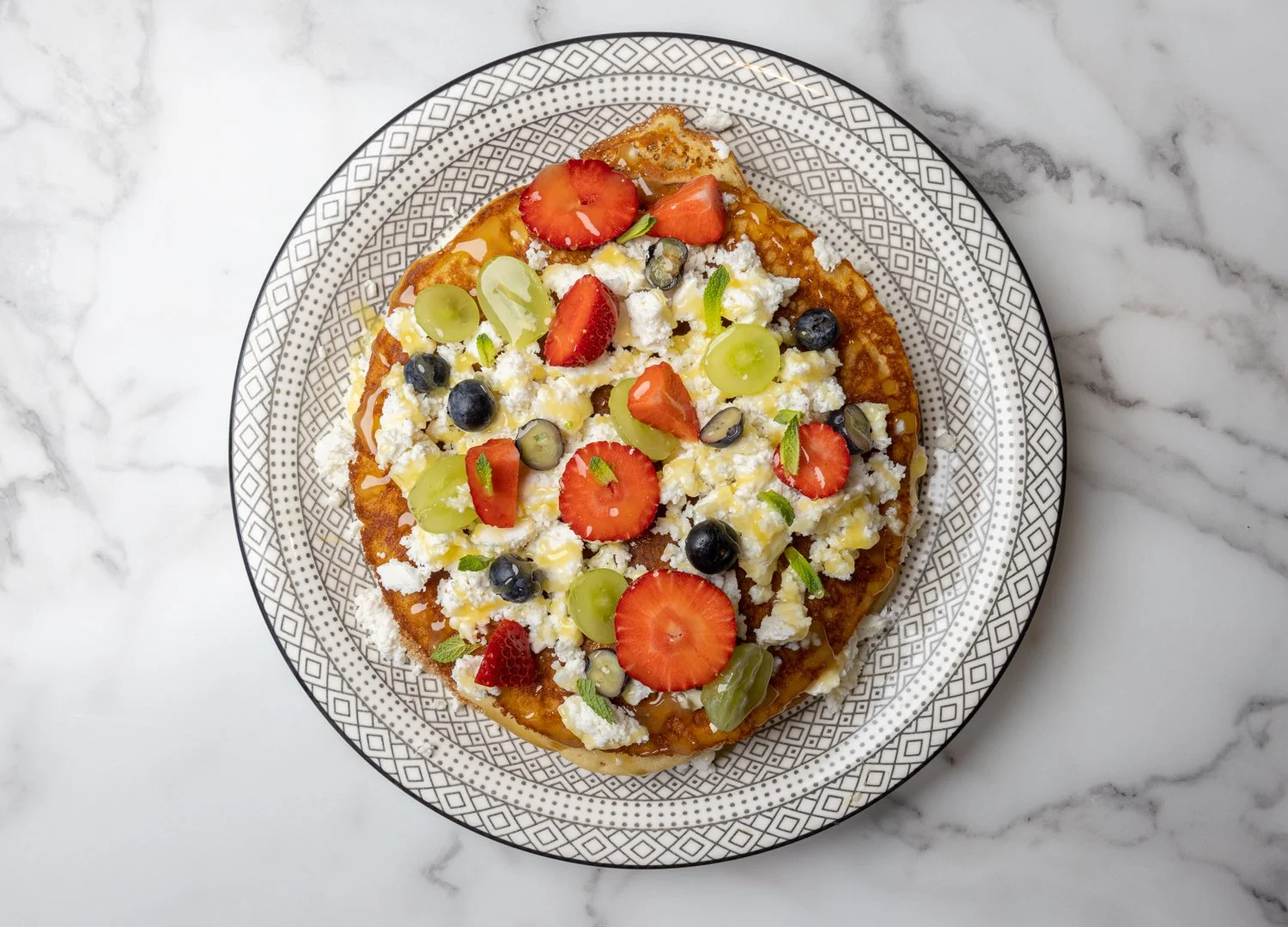 Dessert topped with sliced strawberries, blueberries, green grapes, and crumbled cheese on a decorative plate. Healthy pancakse, brunch, healthy, Nicosia.