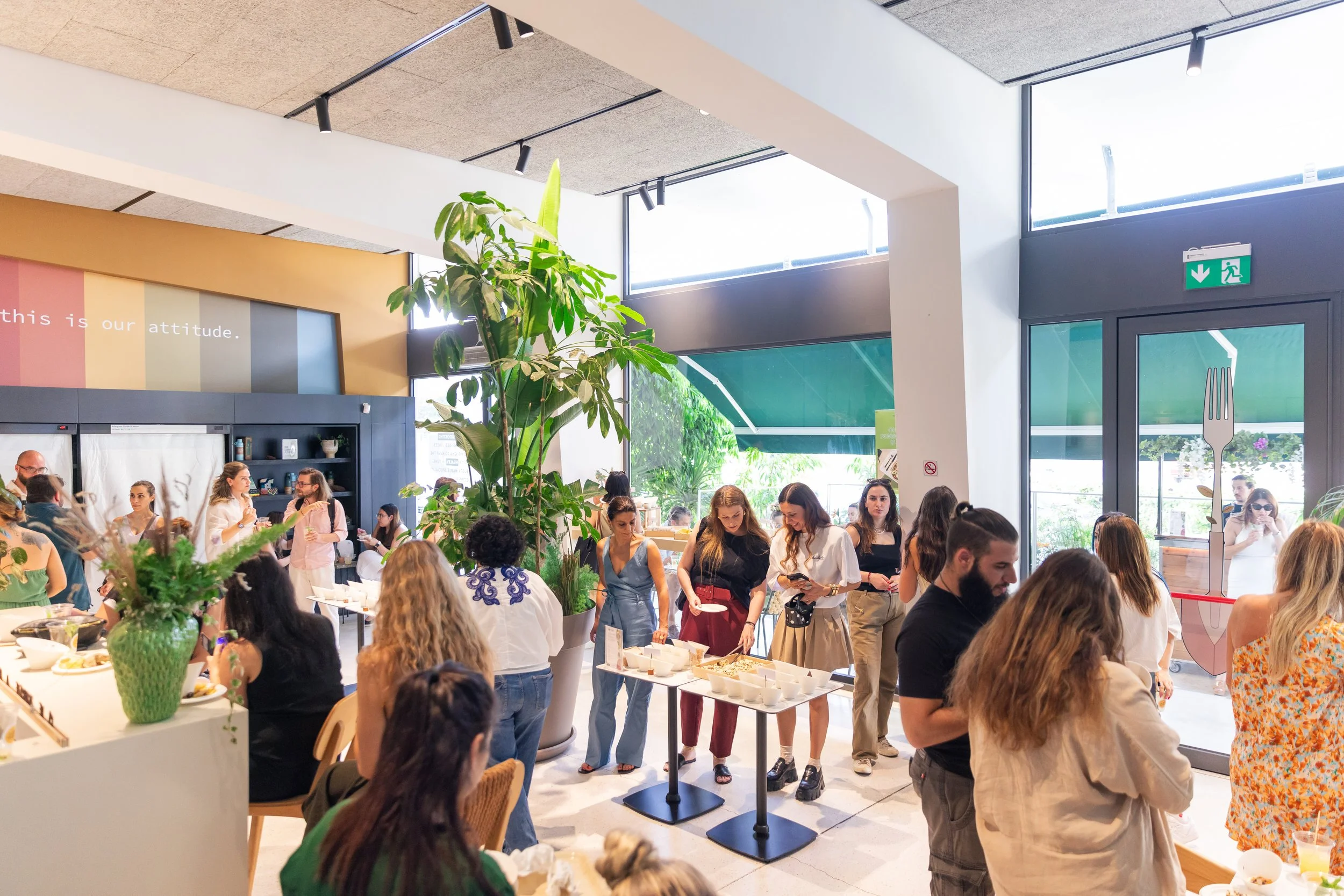 A crowded indoor restaurant or cafe with many people, some standing and others sitting, with large windows letting in natural light, and greenery including a large potted plant in the foreground. Catering, healthy options