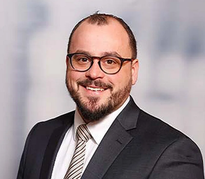 Ein Mann mit Brille, Bart und kurzen Haaren trägt einen Anzug und ein gestreiftes Hemd.